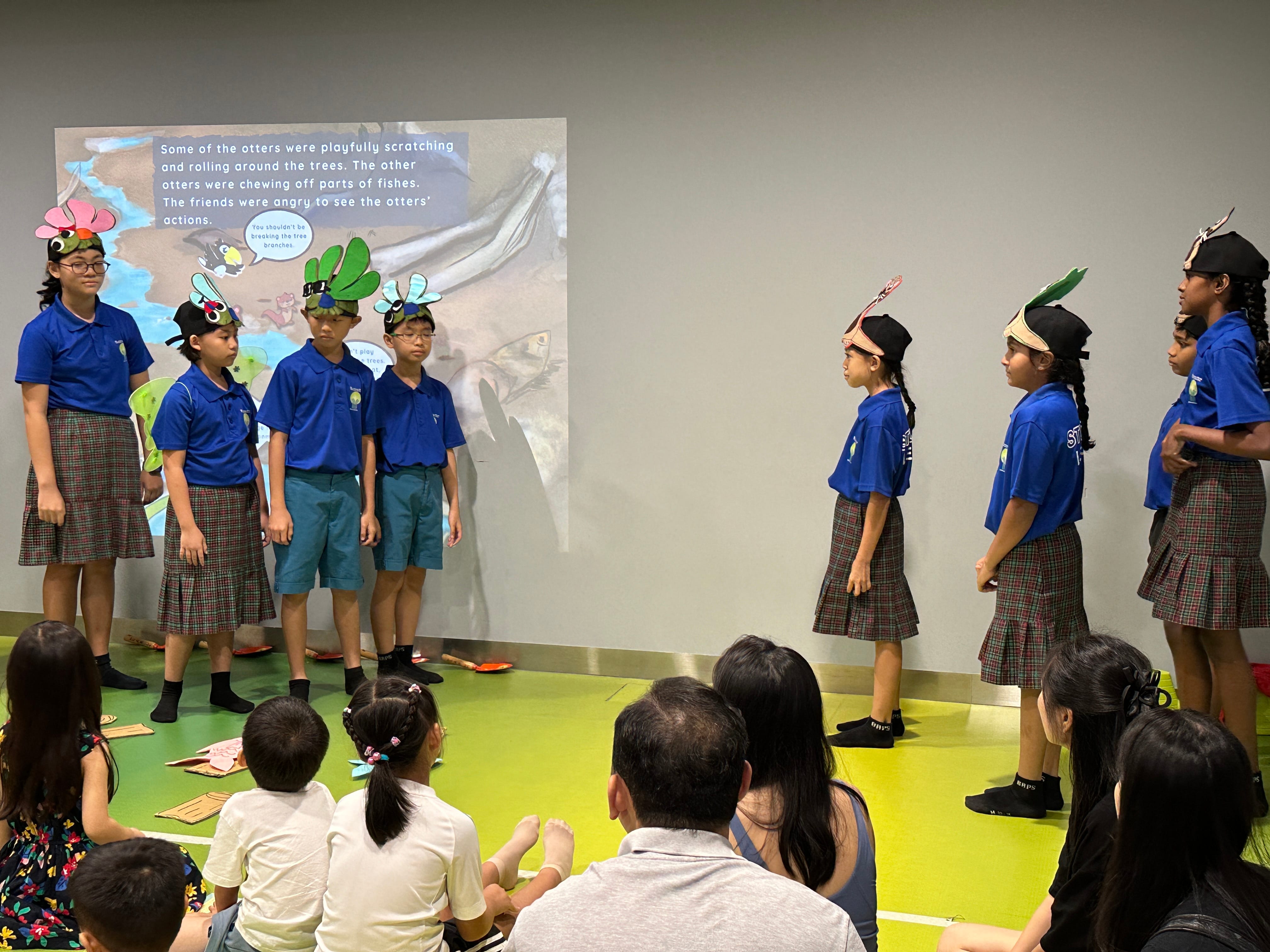 Students in blue shirts and plaid skirts perform in front of a seated audience, wearing animal headpieces.
