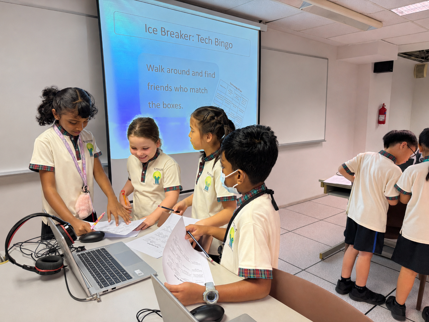 Four children in school uniforms work on worksheets at a desk; slide behind reads Ice Breaker: Tech Bingo.