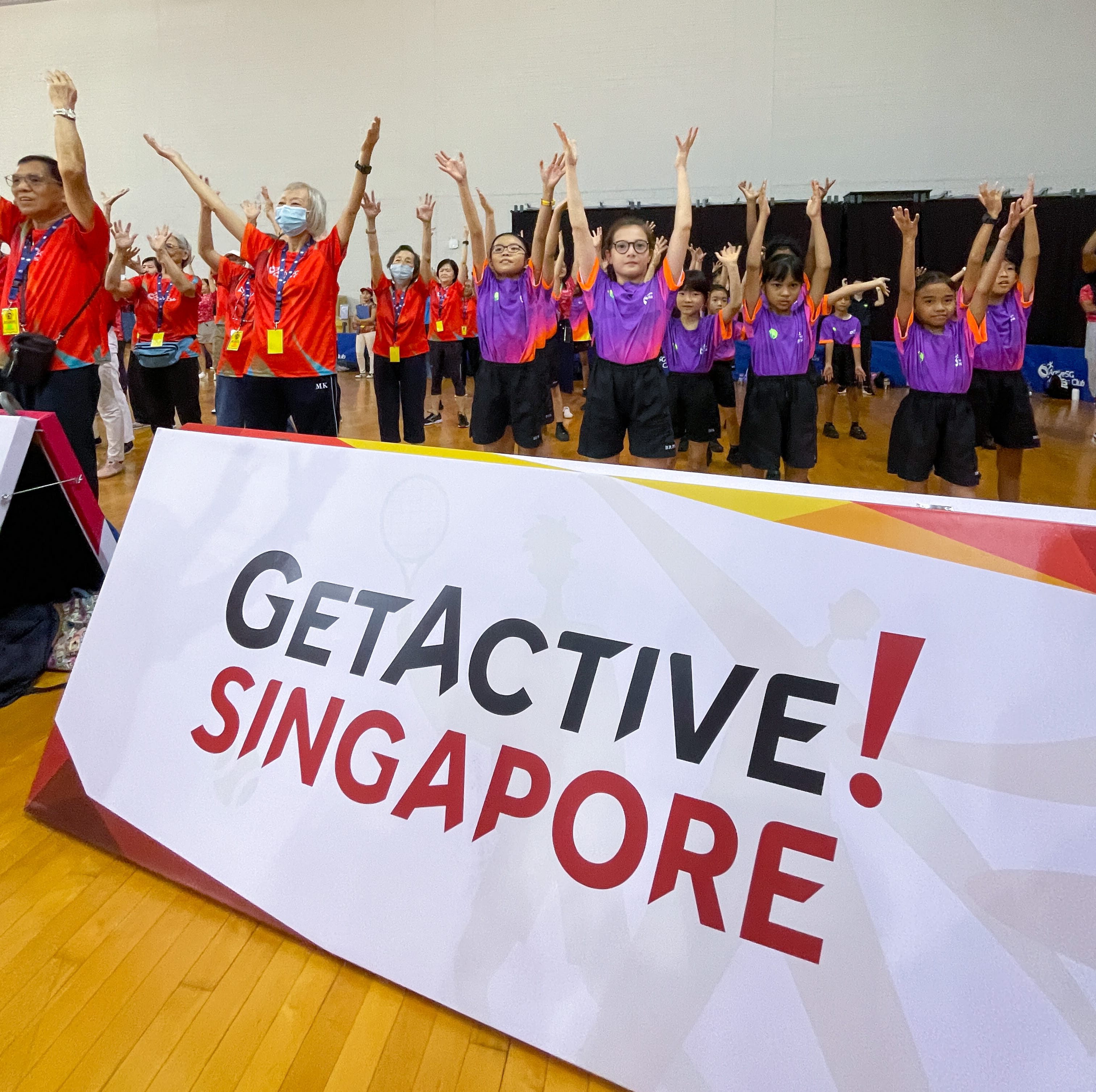 Group in red and purple shirts raising arms in a gym behind a sign reading GetActive! Singapore