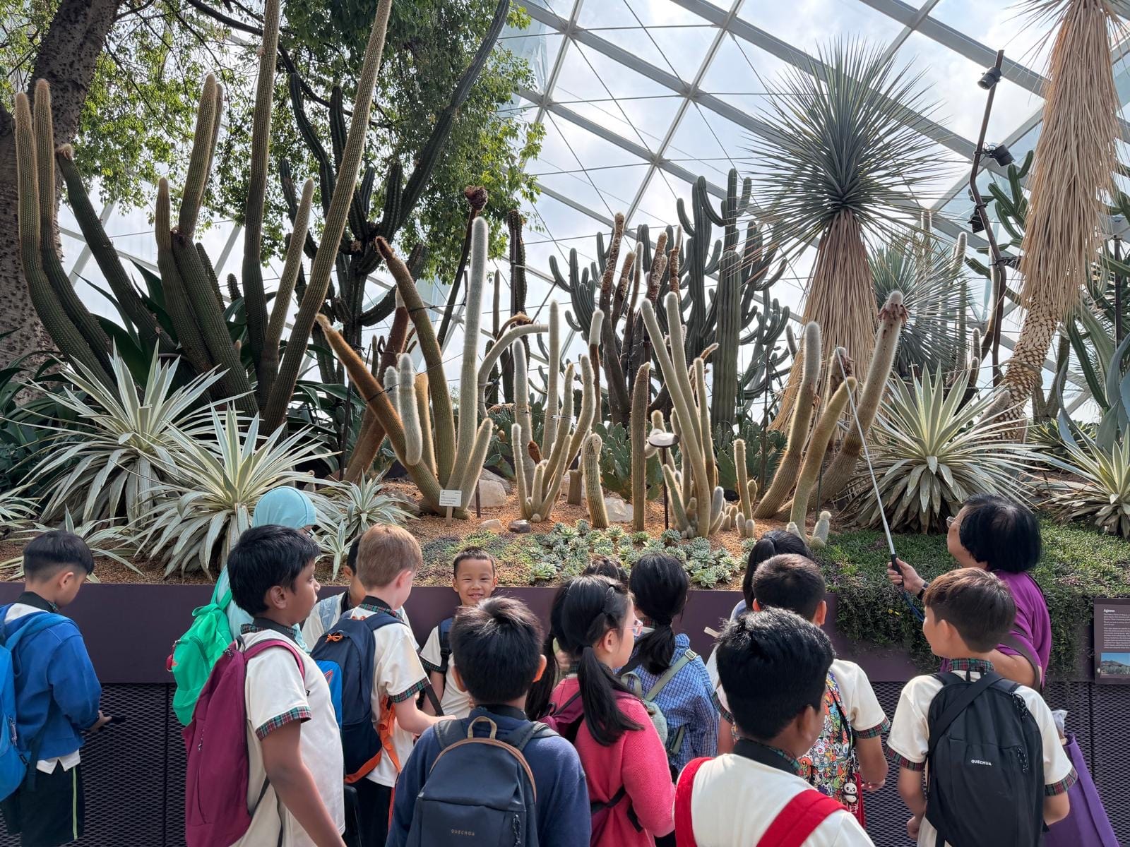Children with backpacks stand in front of a large indoor cactus and succulent display under a glass dome roof.