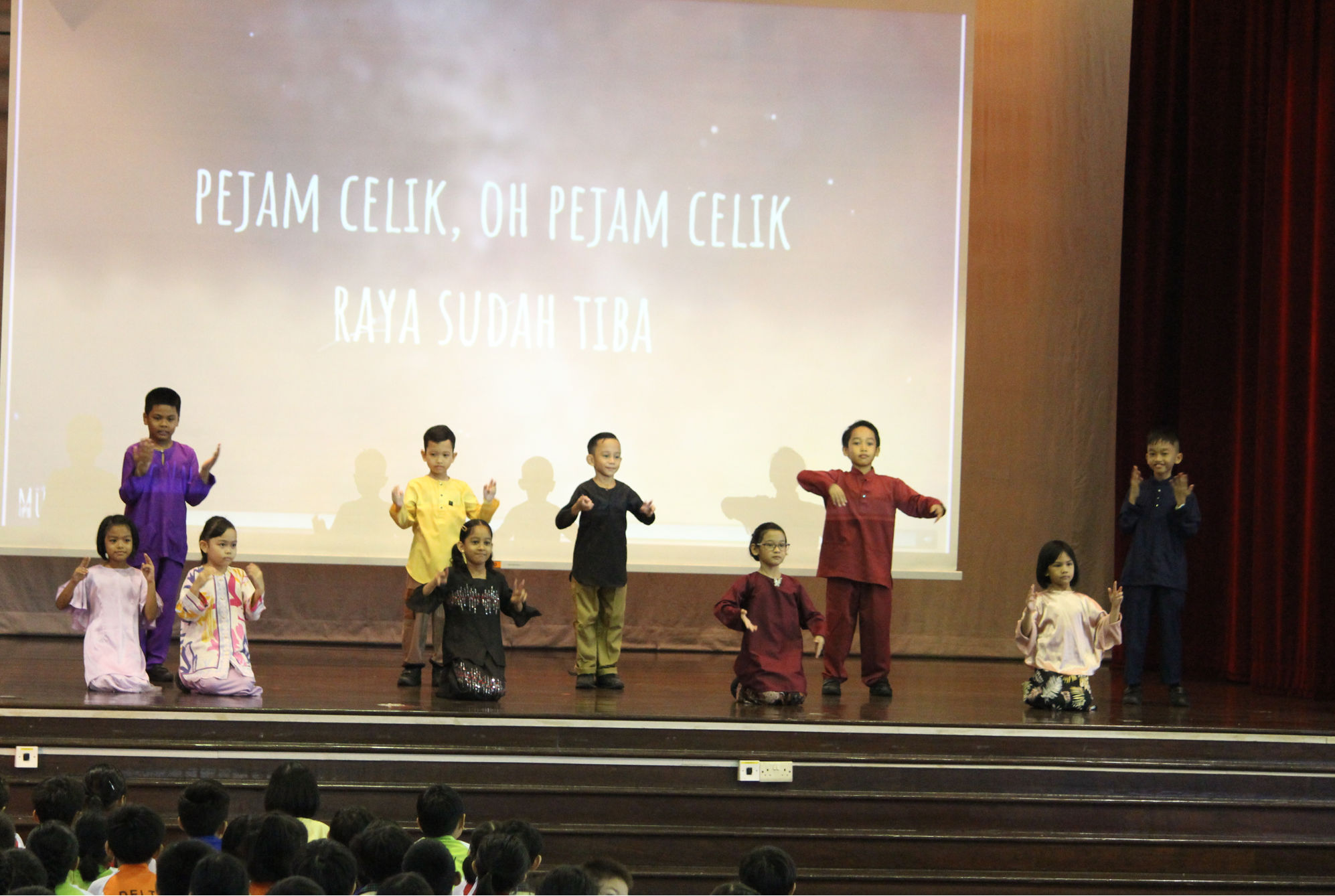 Children in colorful traditional outfits perform on stage before a screen reading Pejam Celik, Raya Sudah Tiba