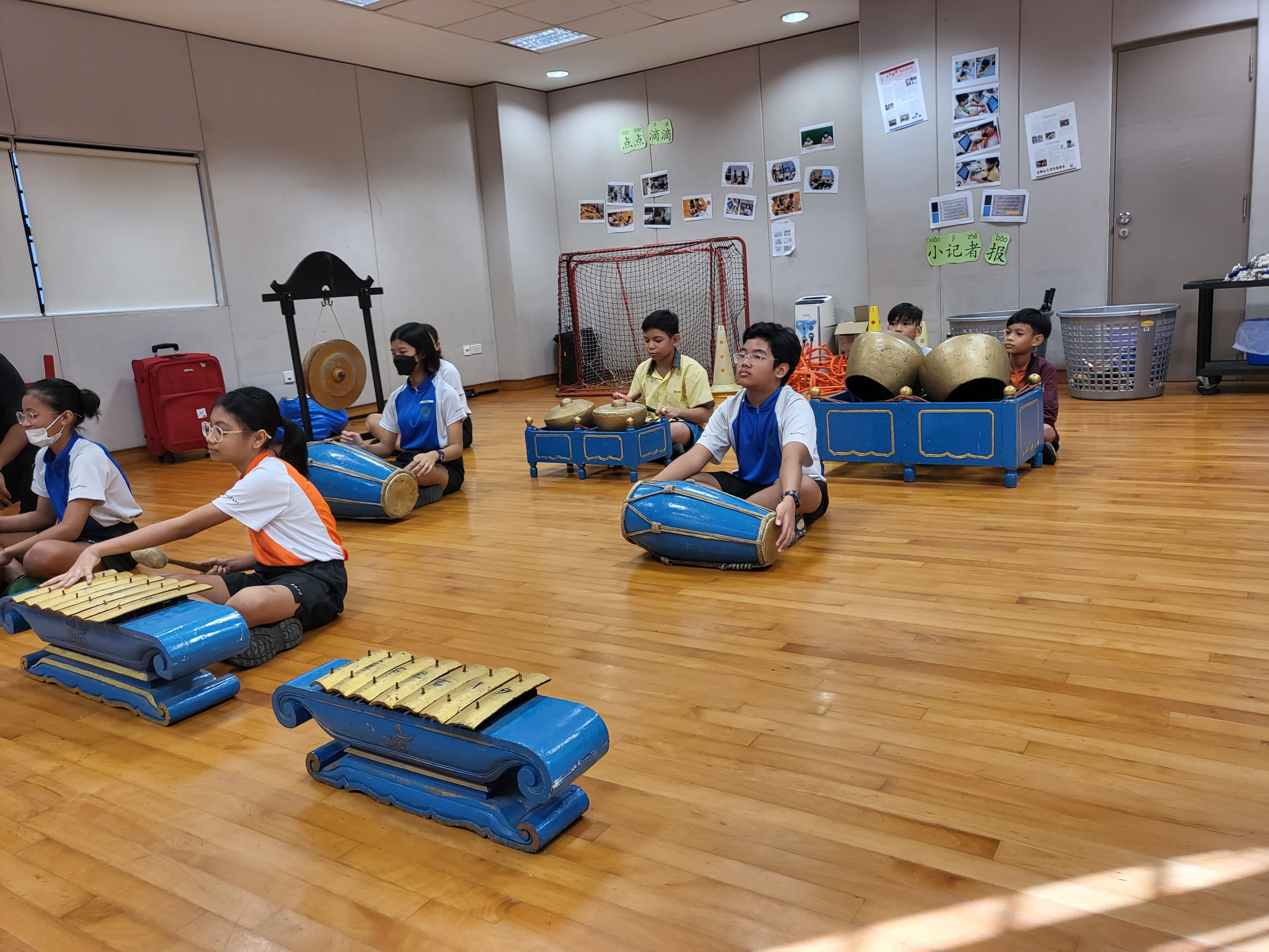 Students sit on a wooden floor playing traditional gamelan instruments with blue decorative stands in a classroom.