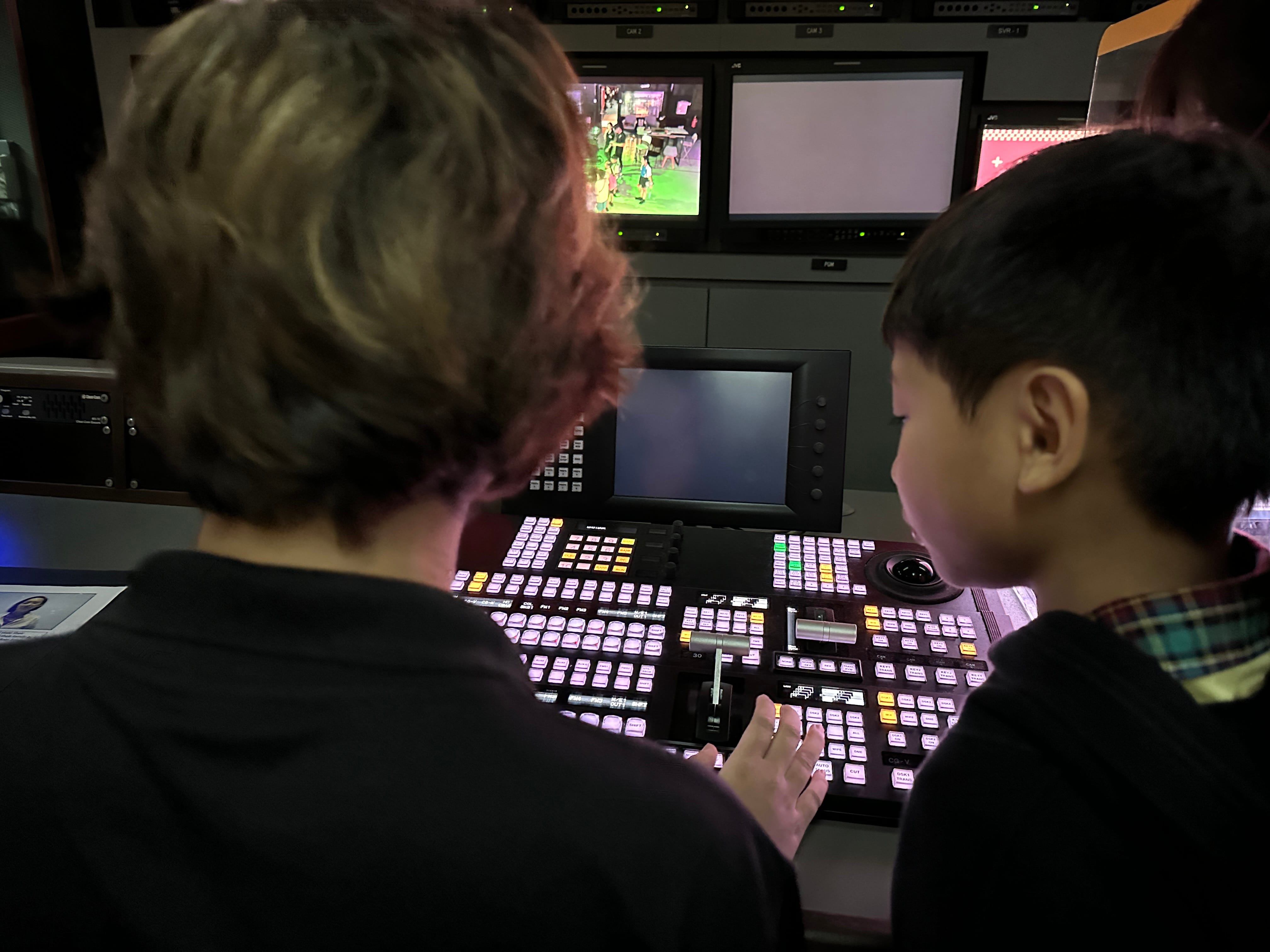 Two children seen from behind operating a large broadcast control panel with multiple monitors displaying video feeds.