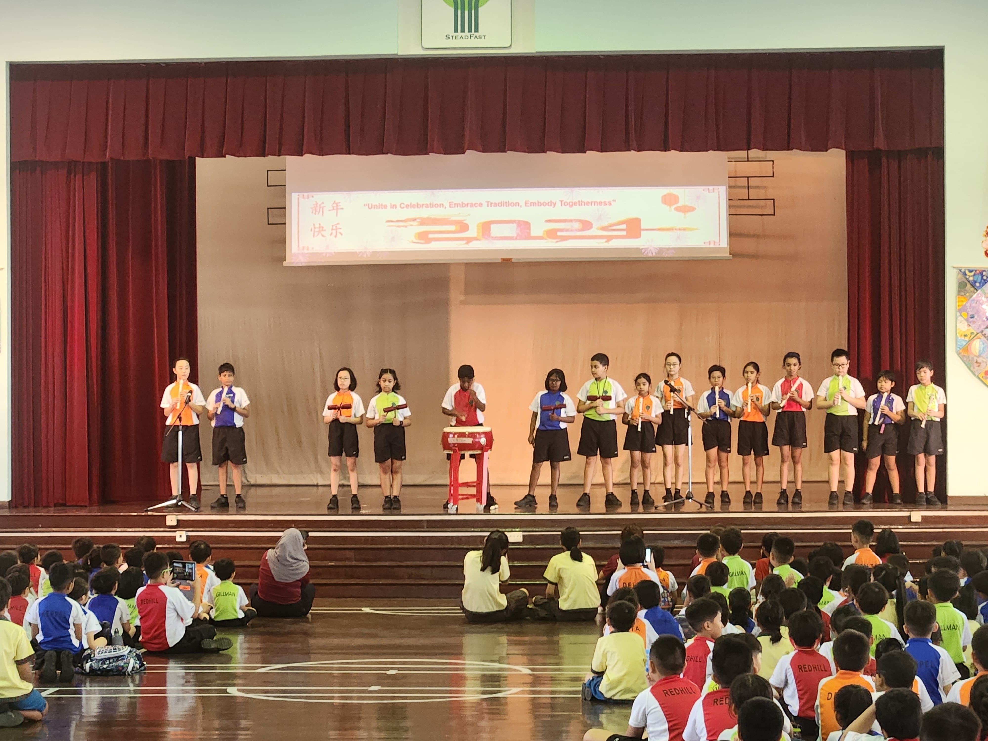 Students perform on stage with instruments at a 2024 Chinese New Year school celebration while seated students watch.