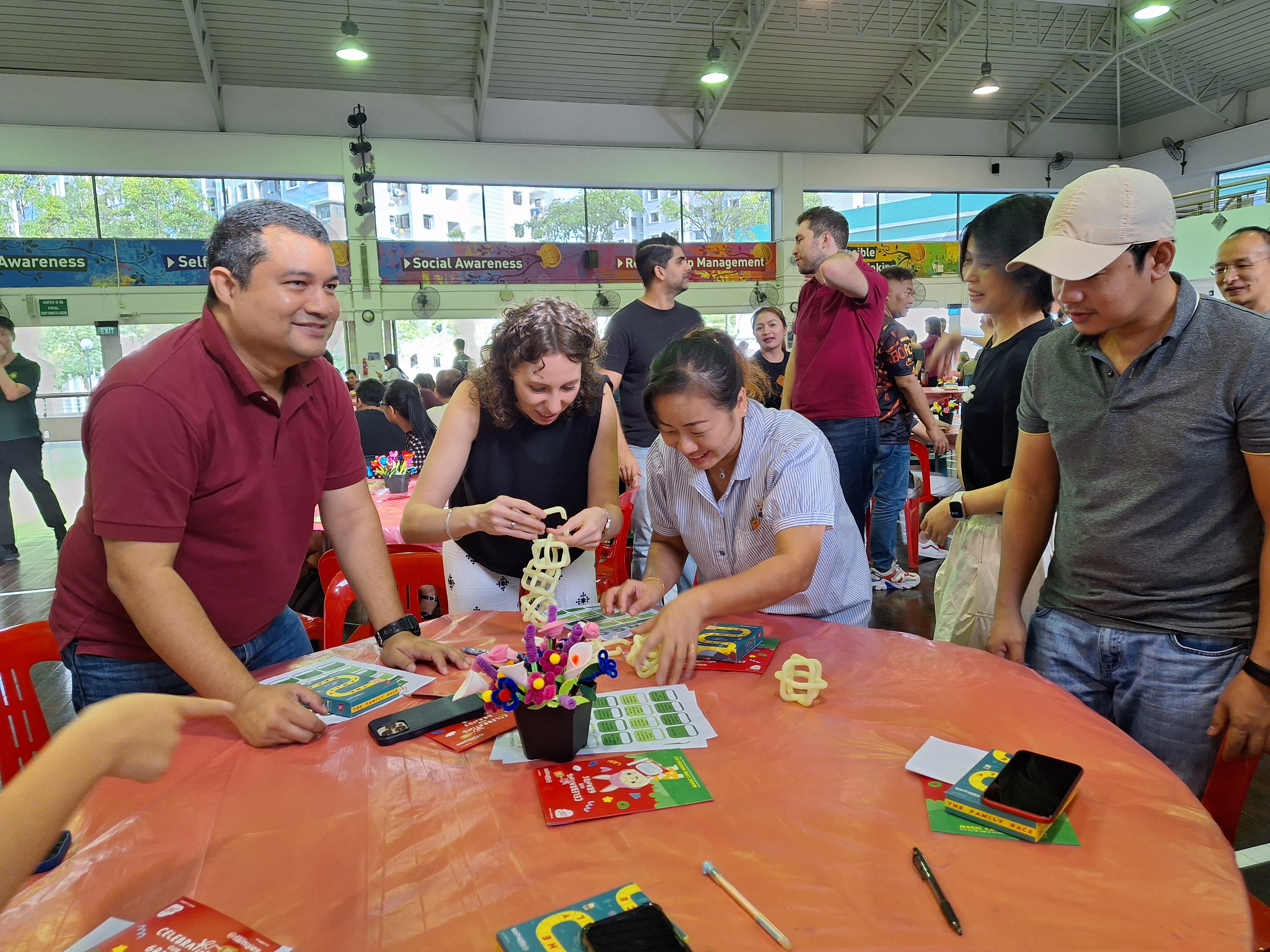 Group stands around a table, smiling and building wooden puzzles in a bright hall with windows.