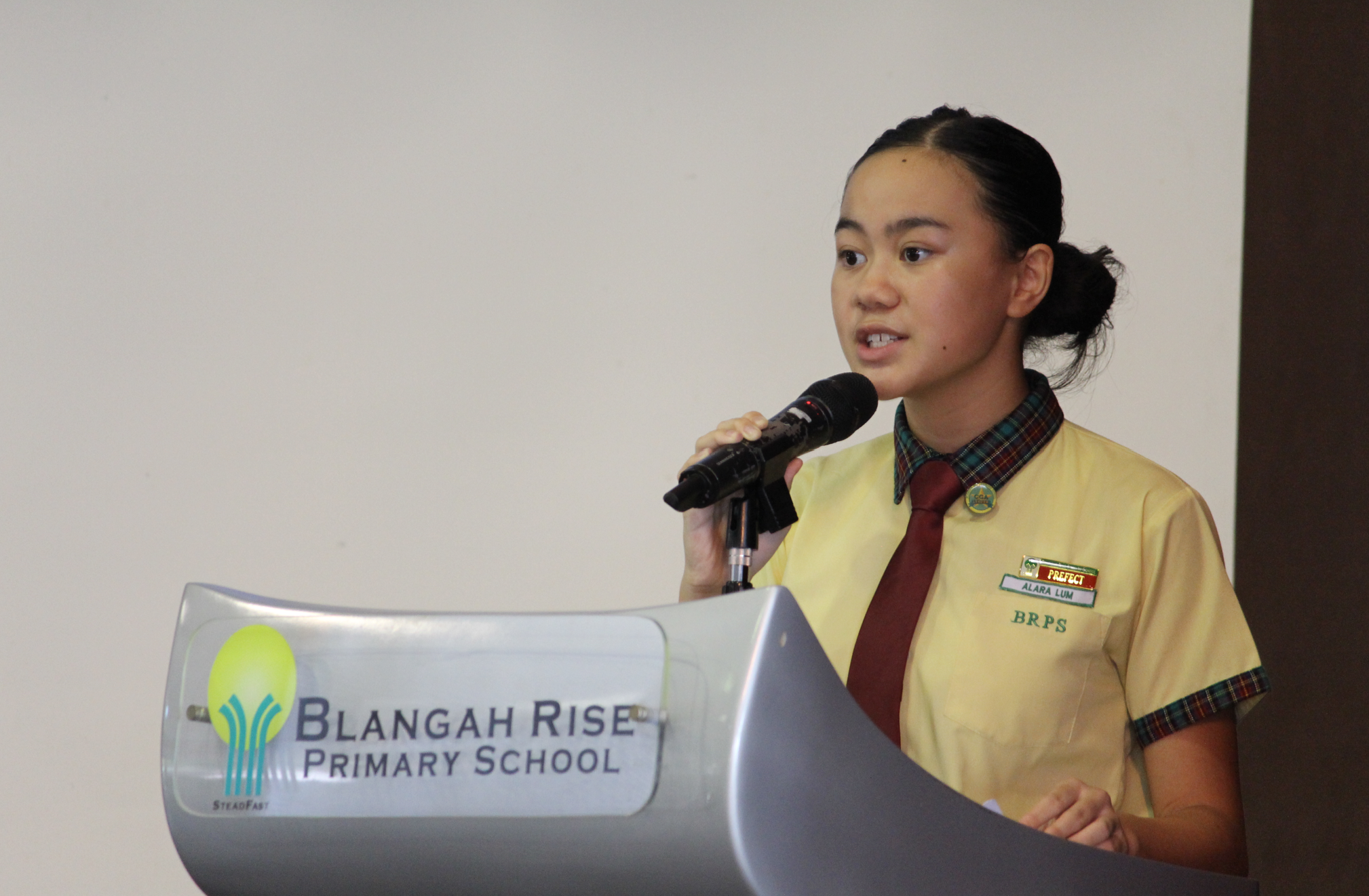 A girl in a yellow BRPS uniform speaks at a Blangah Rise Primary School podium holding a microphone.