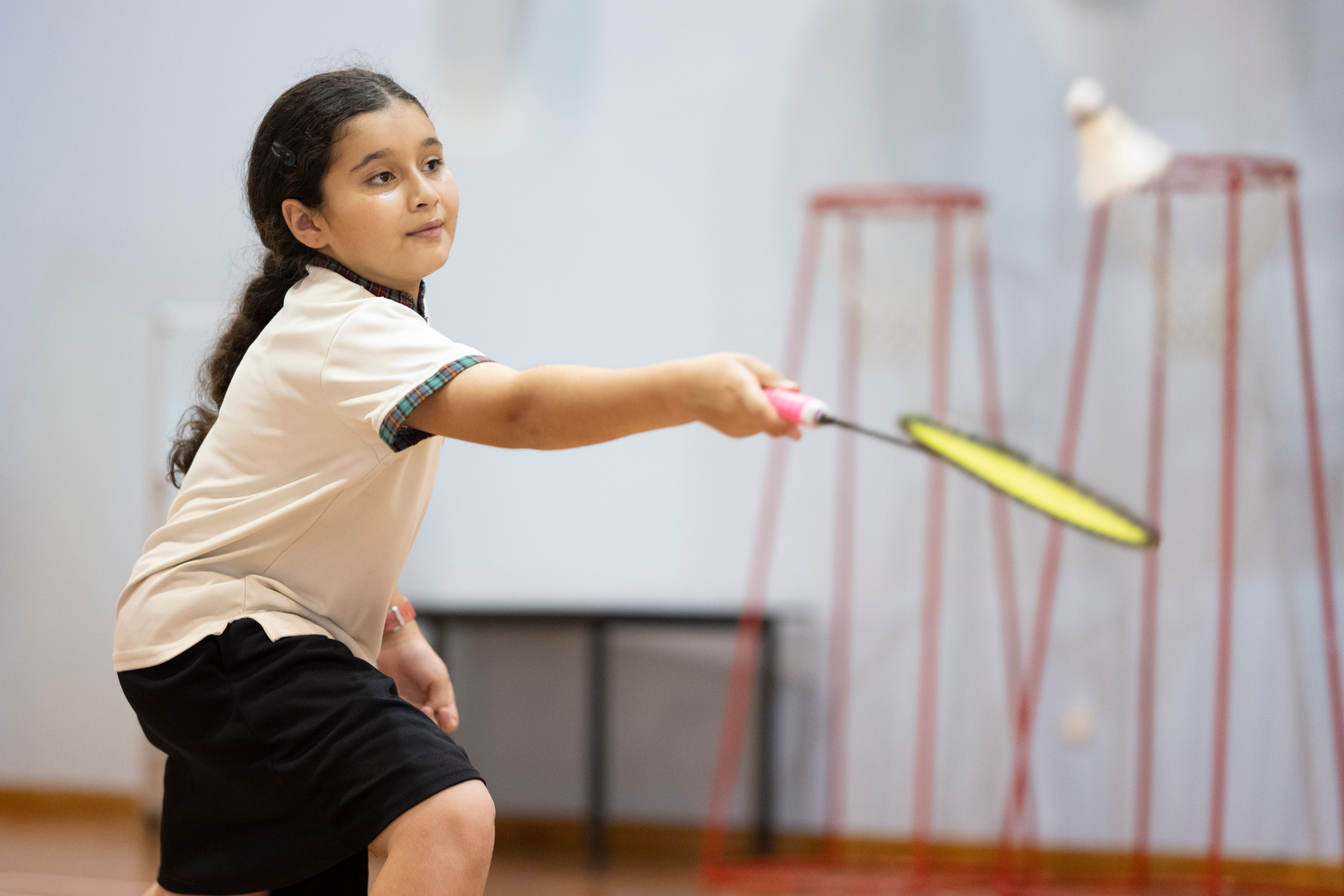 Girl in a school uniform swings a badminton racket in an indoor gym with nets in the background.