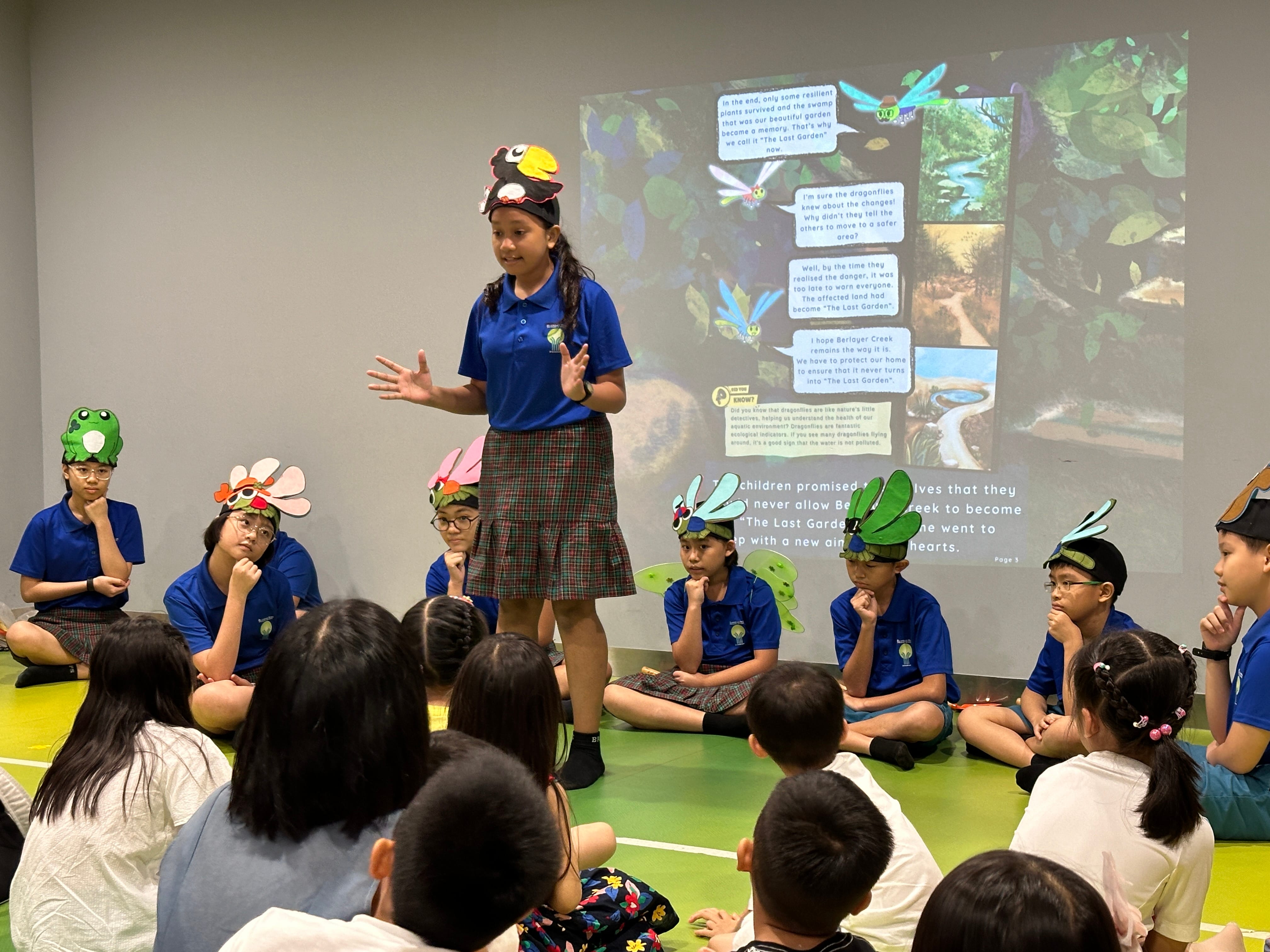 Girl in blue uniform presents to seated classmates in animal hats, with a nature slide projected behind.