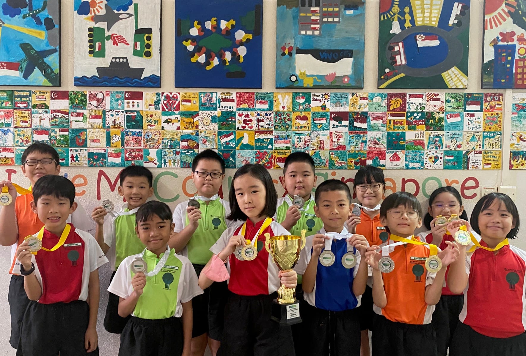 A group of young students in school uniforms holding medals and a gold trophy in front of colorful painted artwork.