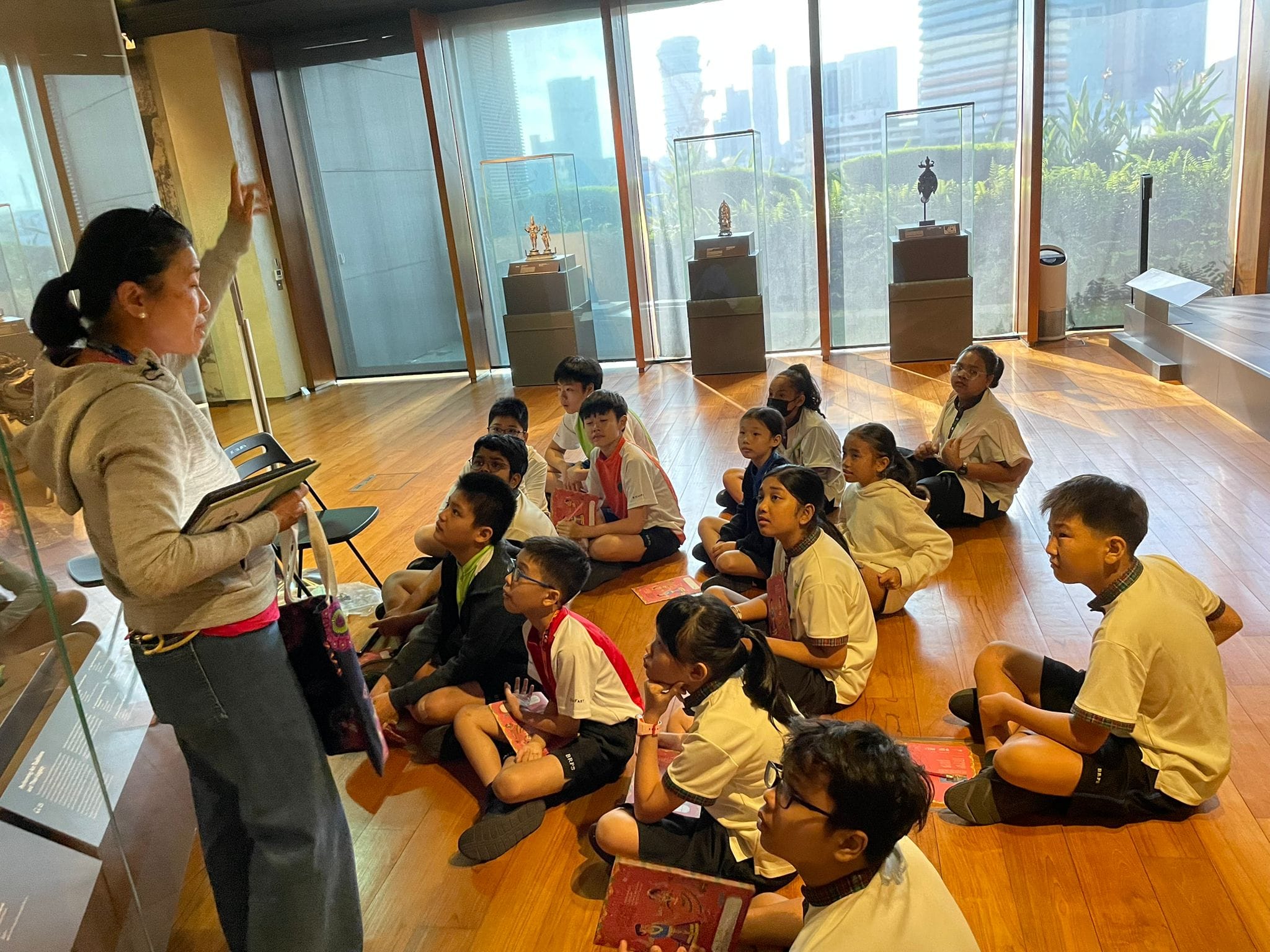 A woman gestures while speaking to a group of children seated on a museum floor, with display cases and city views behind them.