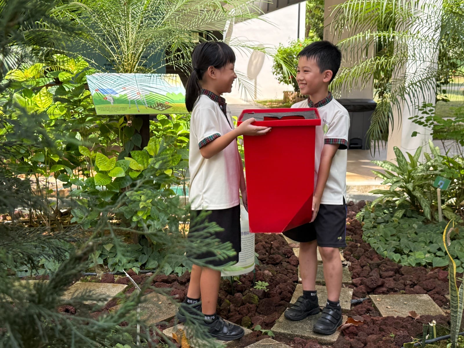 Two children in school uniforms hold a red bin together while smiling at each other in a lush garden.