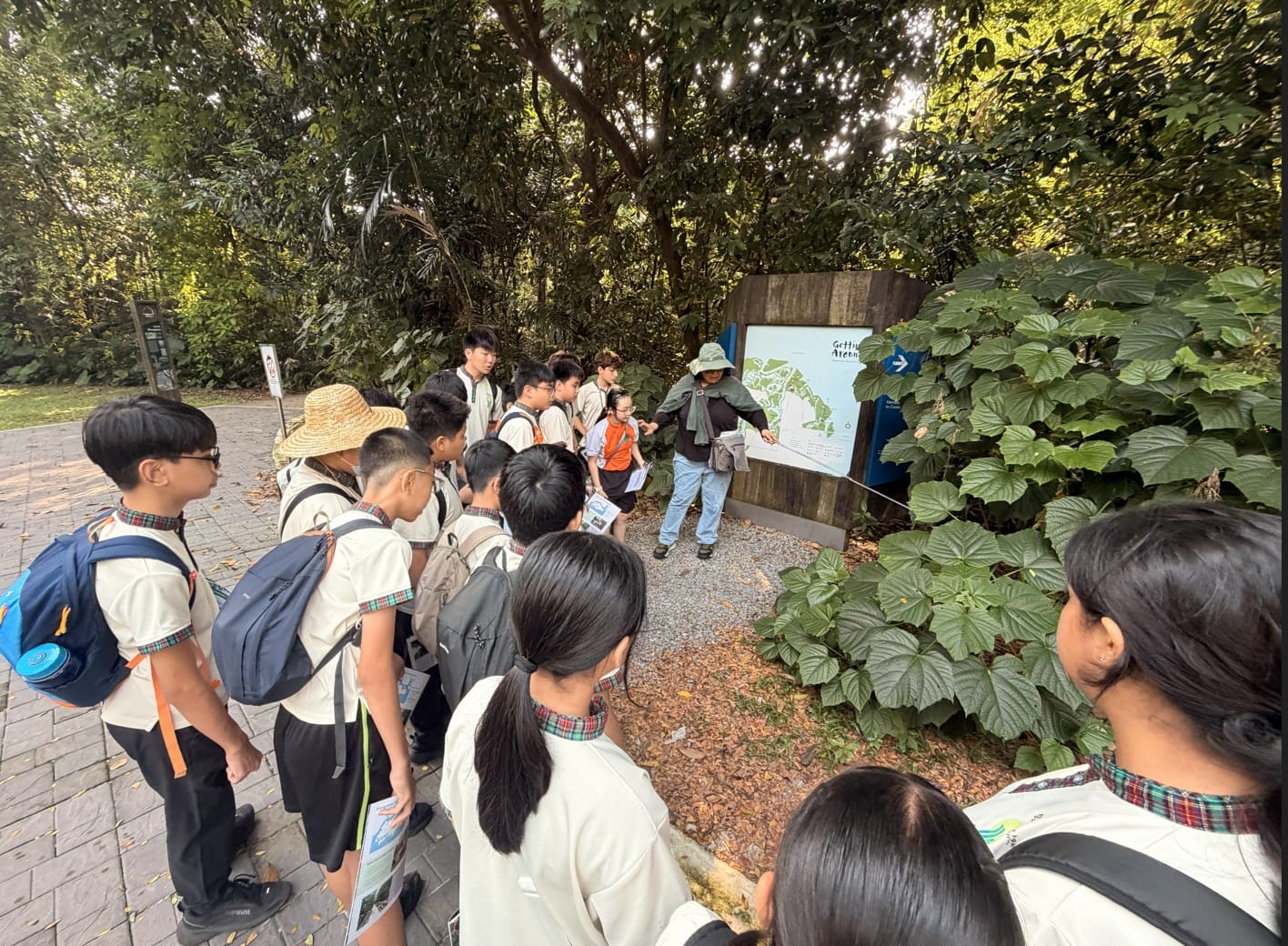 A guide in a hat points at a park map board while uniformed students with backpacks gather around in a forest setting.