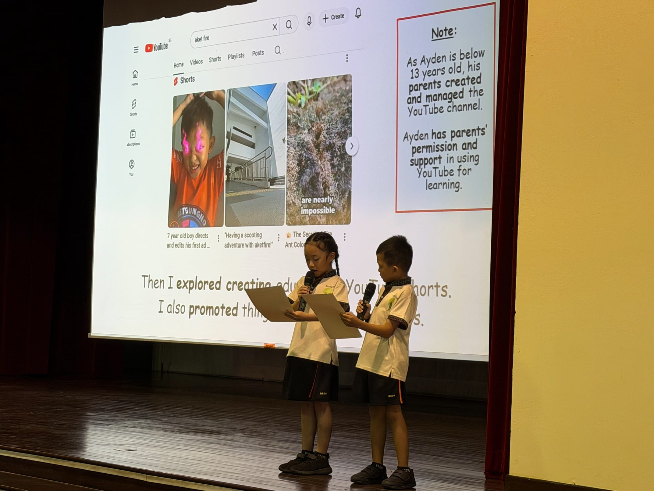 Two young students in school uniforms present on stage with microphones in front of a YouTube-themed slideshow.