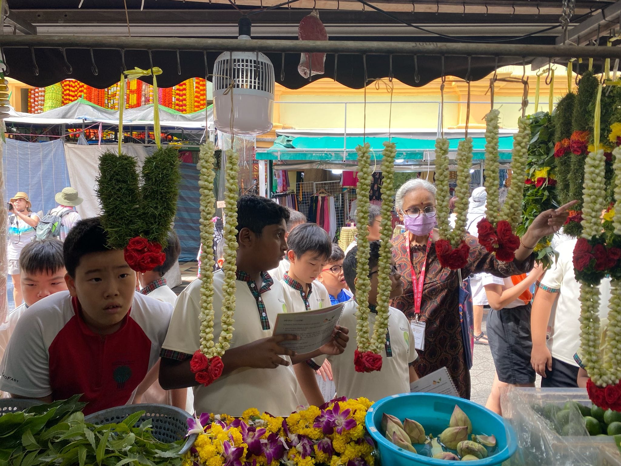 Children and an adult woman browse a flower garland stall at a colorful outdoor market.