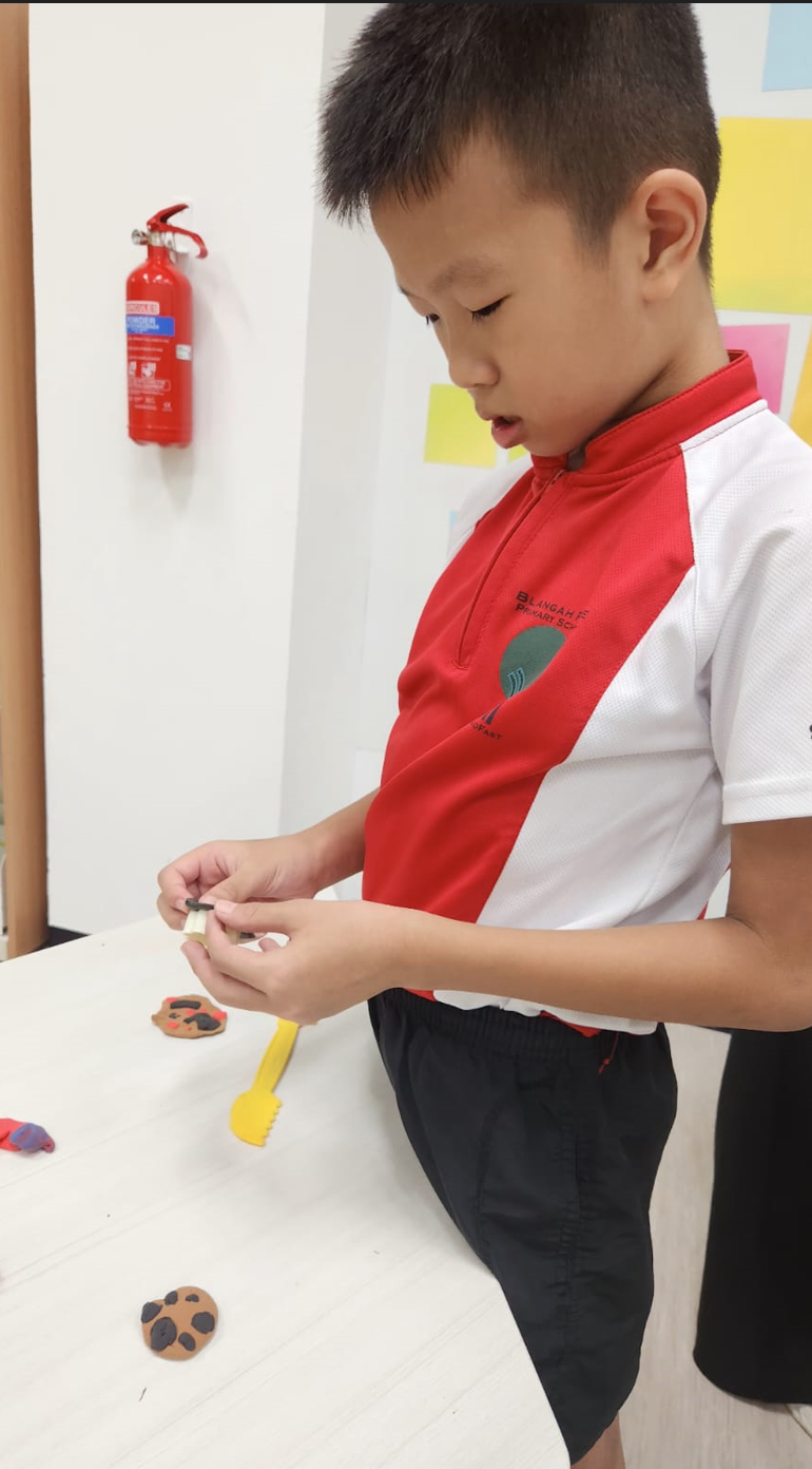 A young boy in a school uniform works with clay pieces on a white table, with a fire extinguisher on the wall behind.