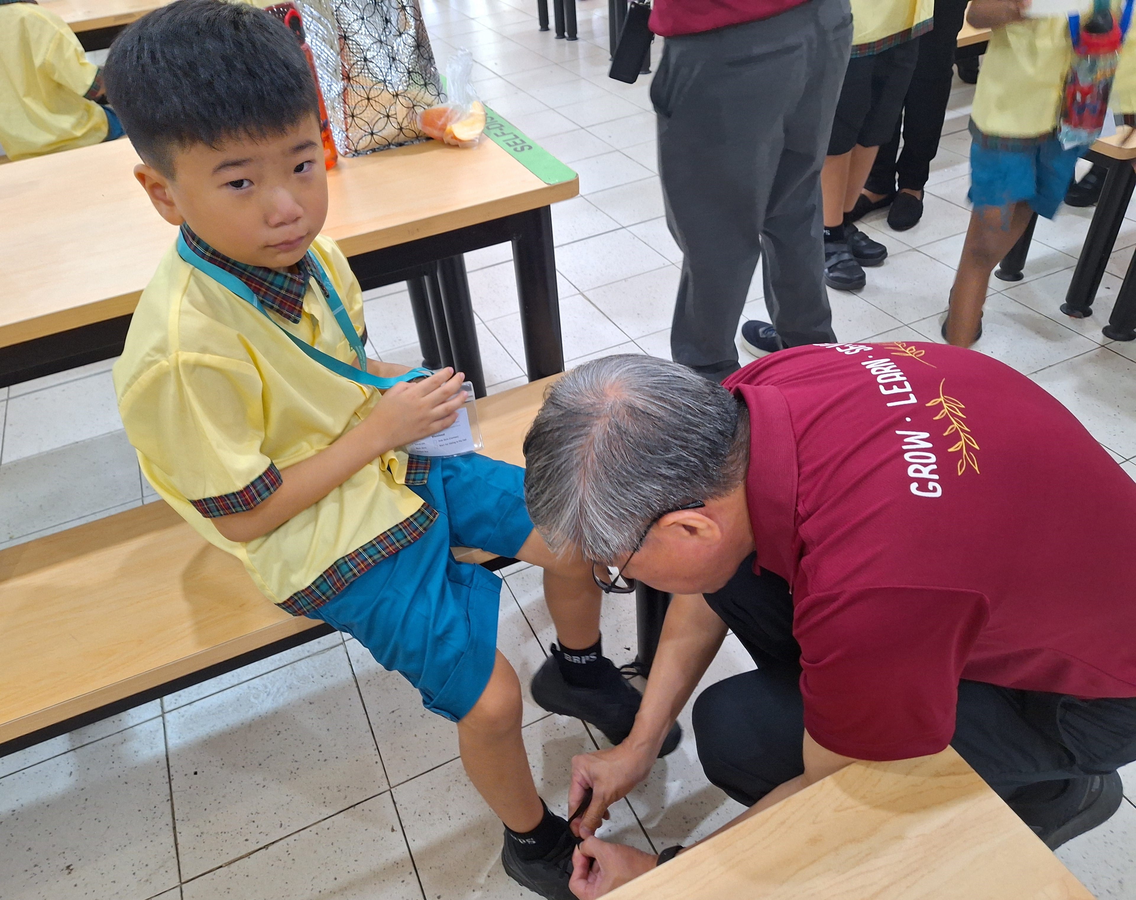 An older man in a red shirt kneels to tie the shoe of a young boy in a yellow school uniform.