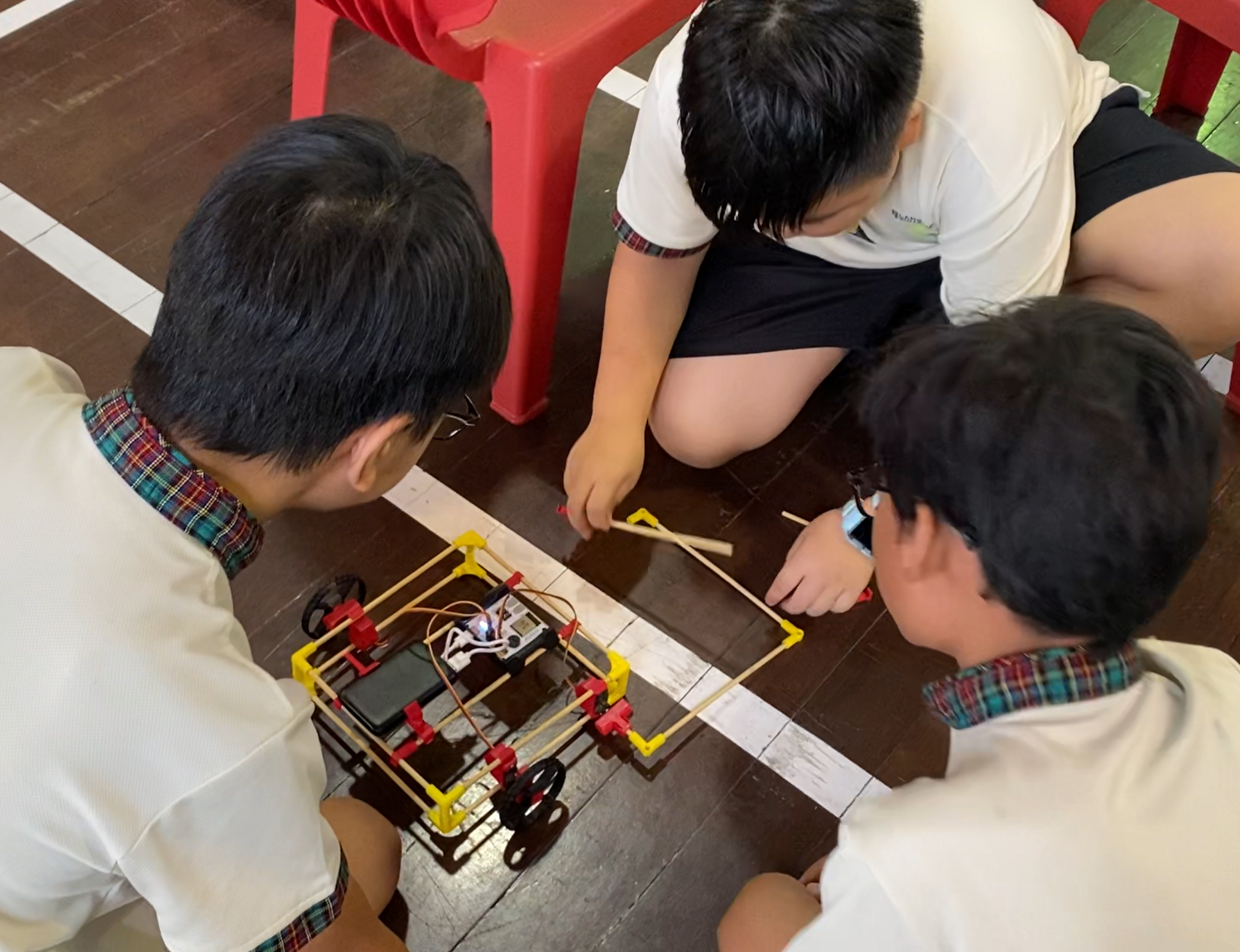 Three students kneel on a gym floor, assembling a small wheeled robot with sticks and wires.
