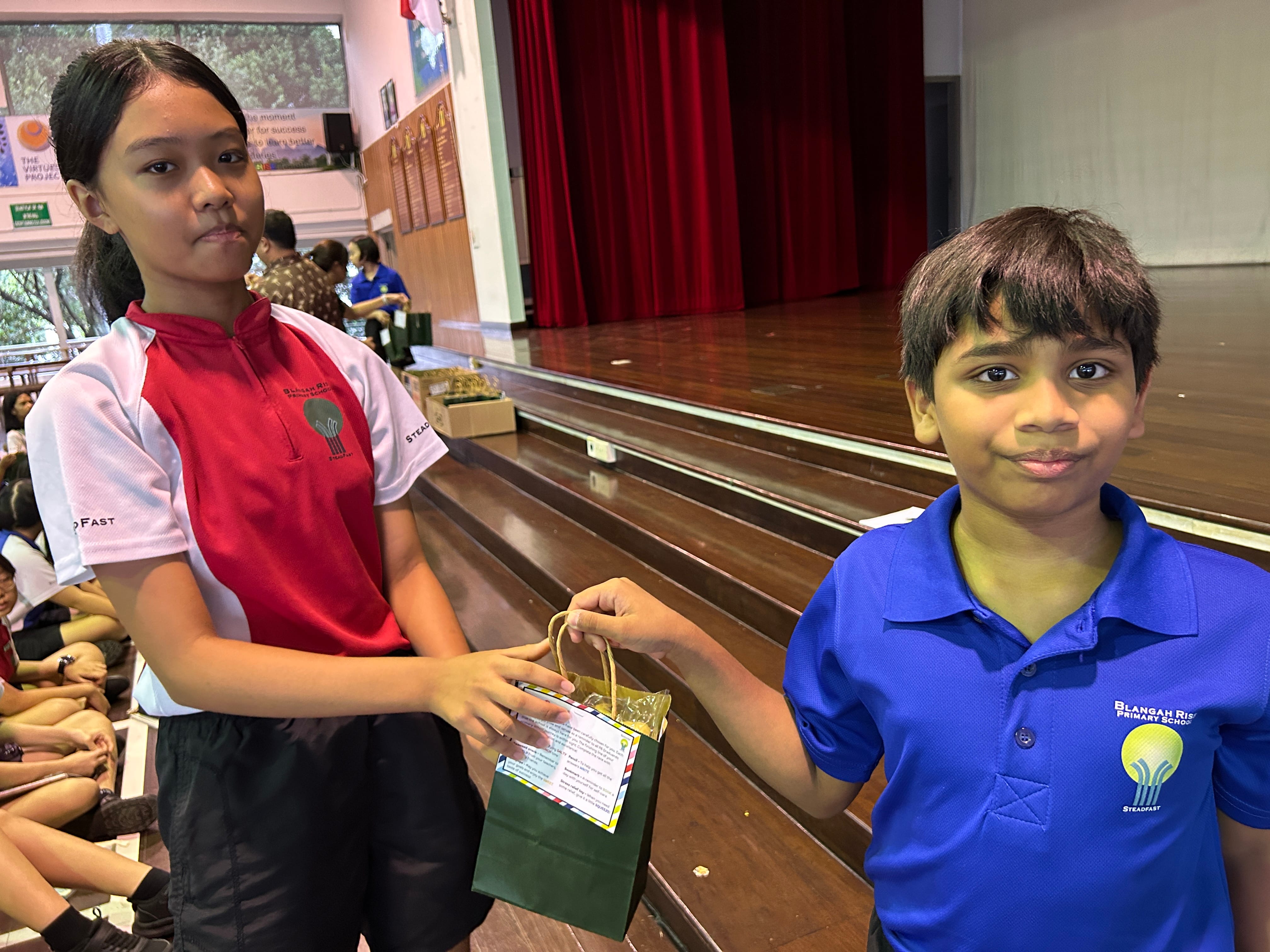 Two school children in uniforms hold a small green gift bag together in a school hall with a wooden stage.