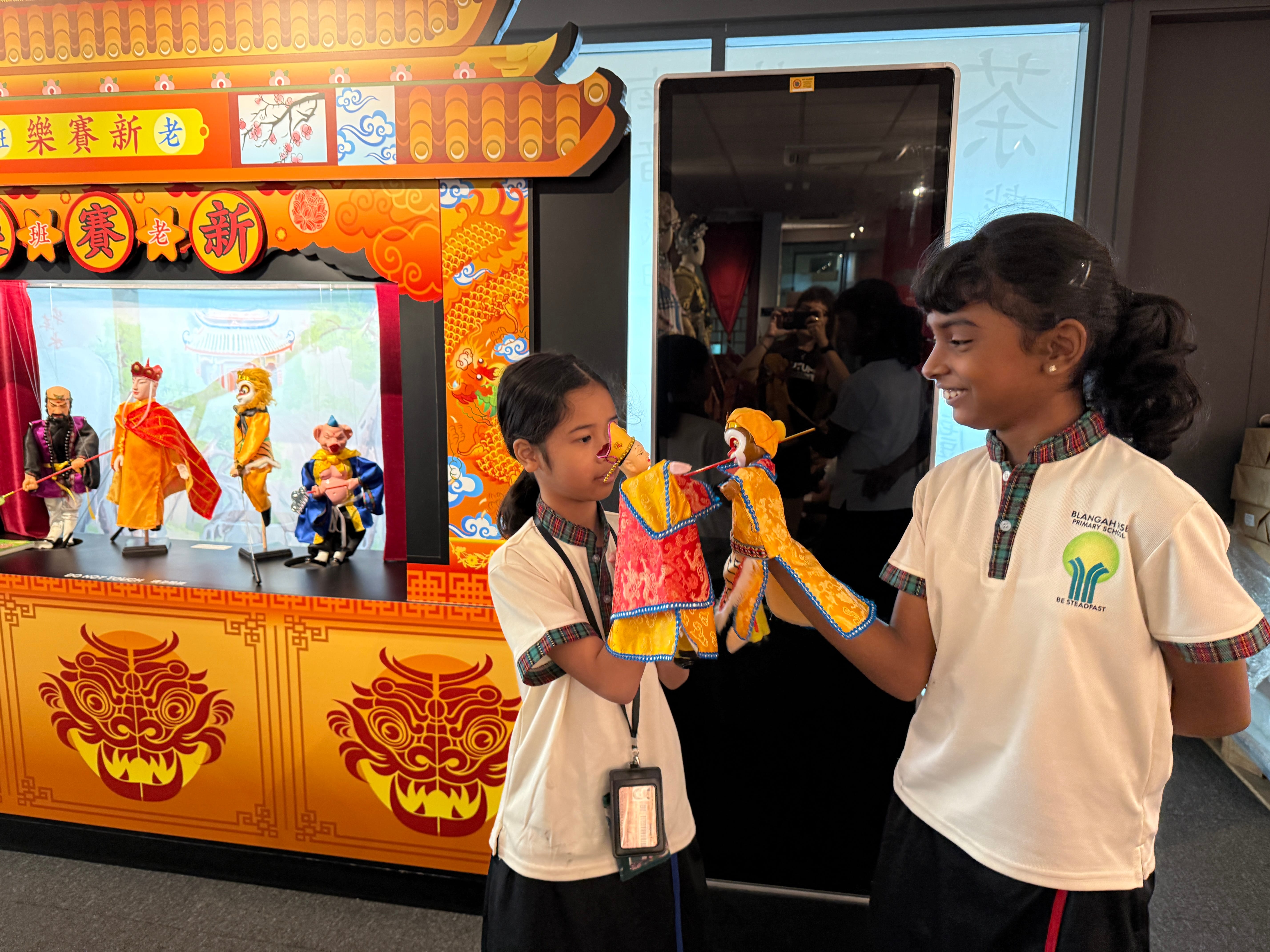Two schoolgirls hold colorful puppets in front of a bright orange traditional puppet stage display.