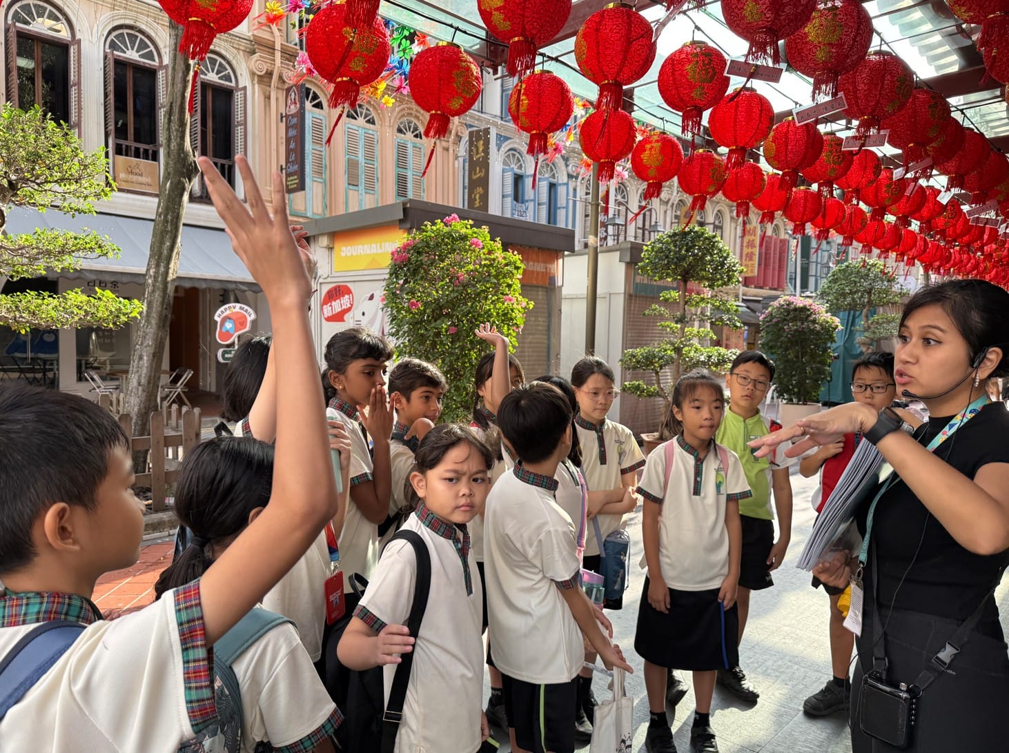 A tour guide speaks to a group of school children on a street lined with red hanging lanterns and shophouses.