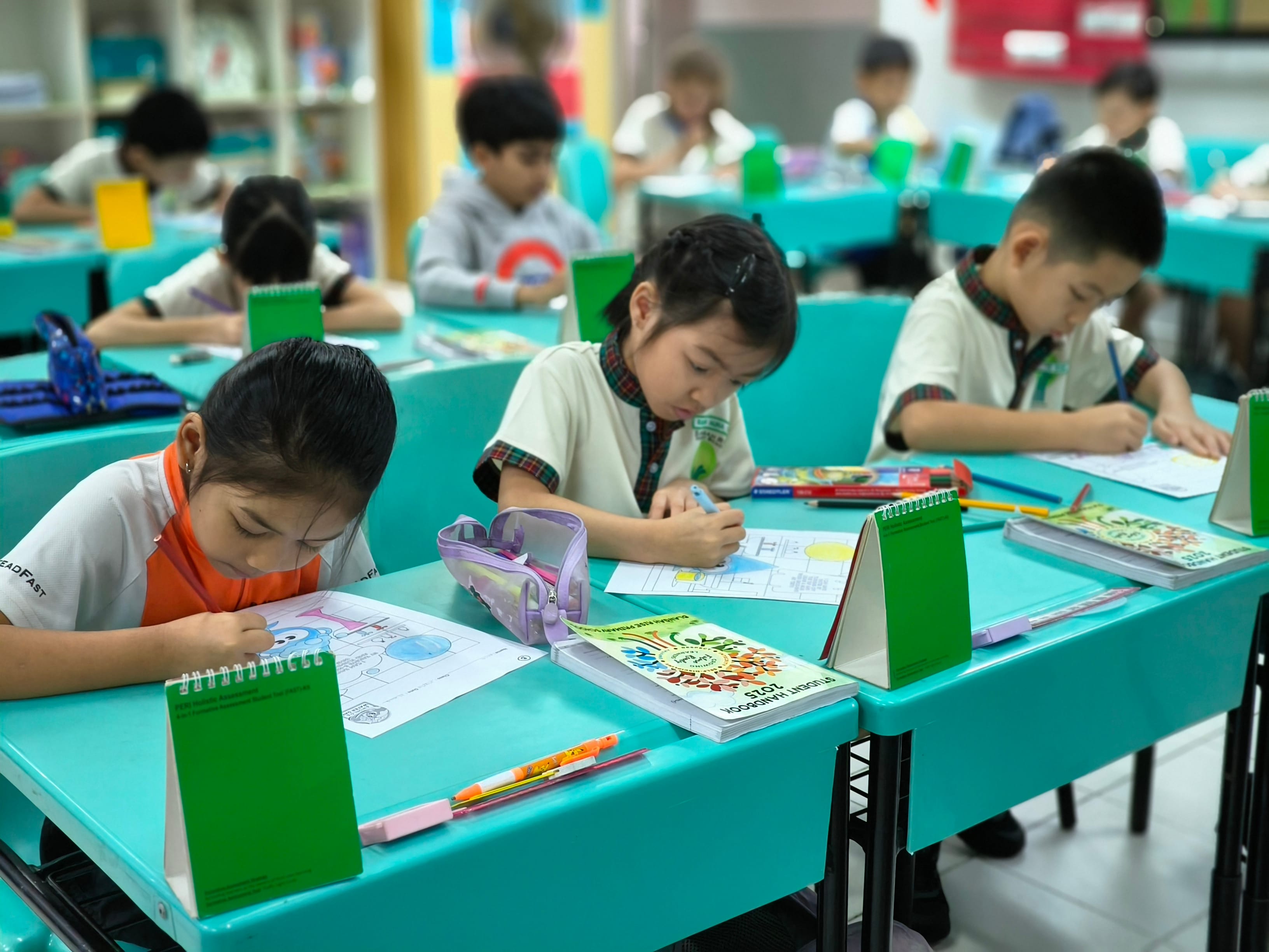 Children in a classroom sit at teal desks, writing and coloring on worksheets with pencils and crayons.
