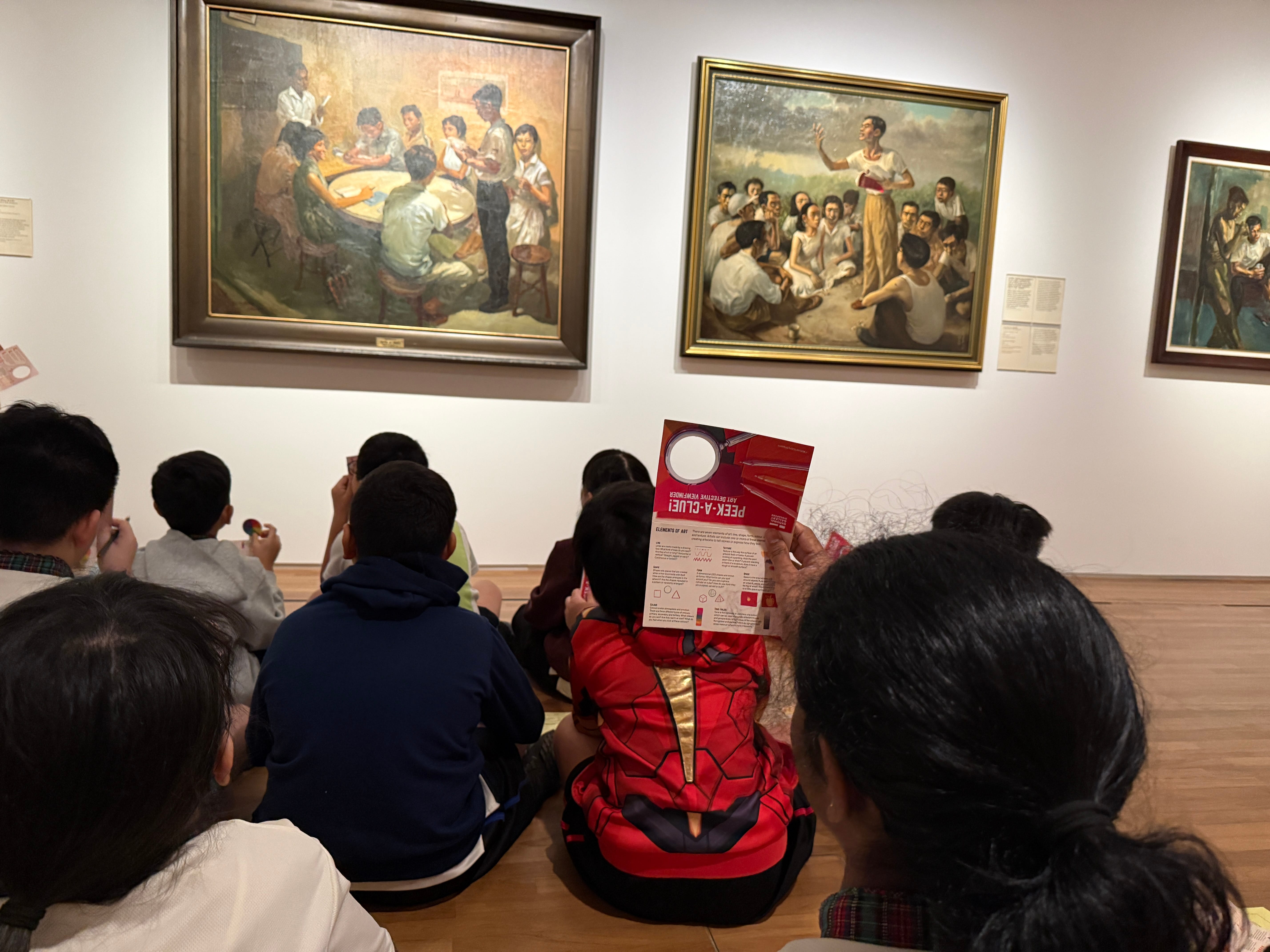 Children sit on a museum floor facing framed oil paintings, one child holds a red Peek-A-Clue activity card.