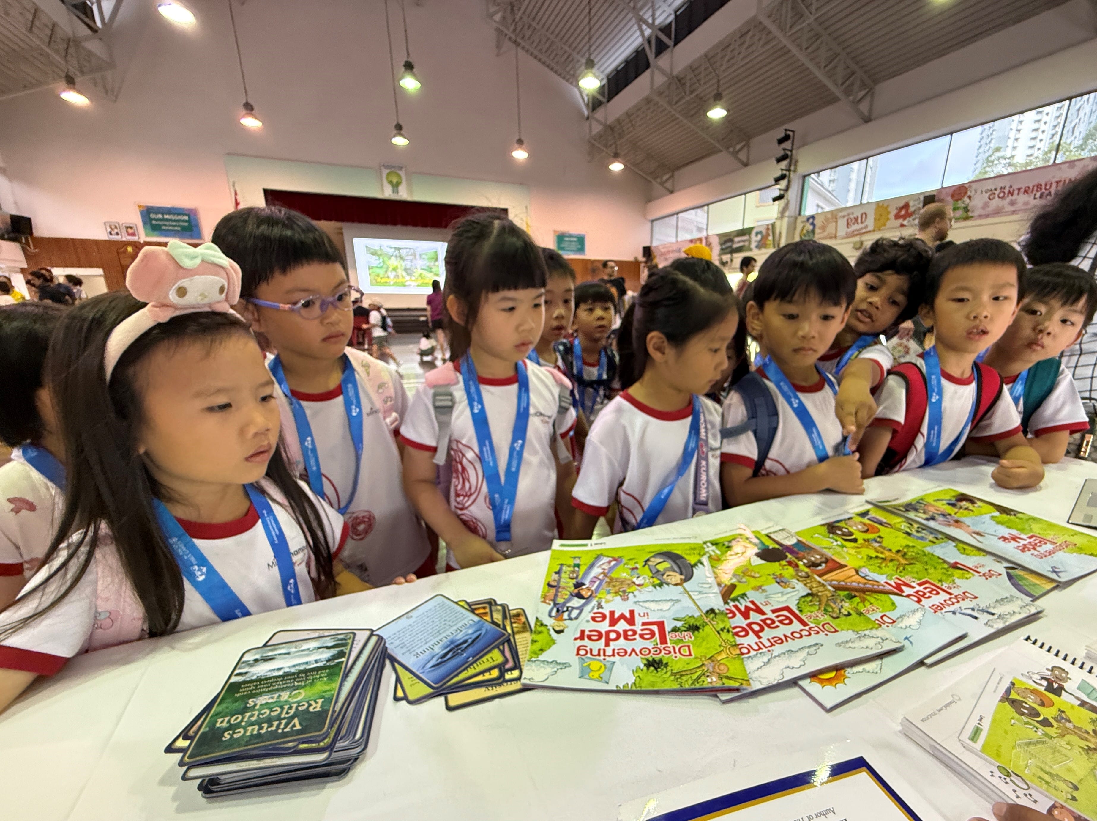 Group of children with lanyards stand at a table, looking at colorful books and card decks in a hall.