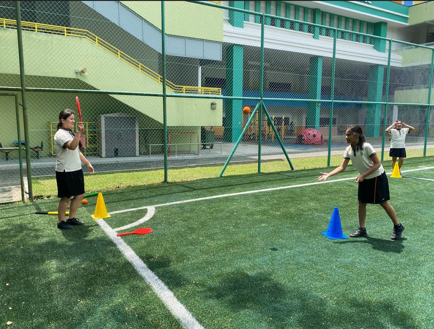 Two school girls play a bat and ball game on a green outdoor court with colorful cones marking the area.