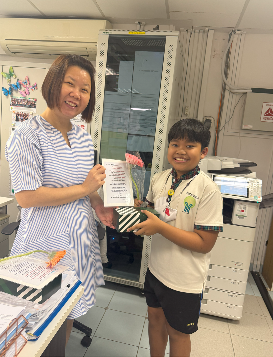 A smiling boy in school uniform presents a woman in a striped dress with flowers, a gift box, and a card in an office.