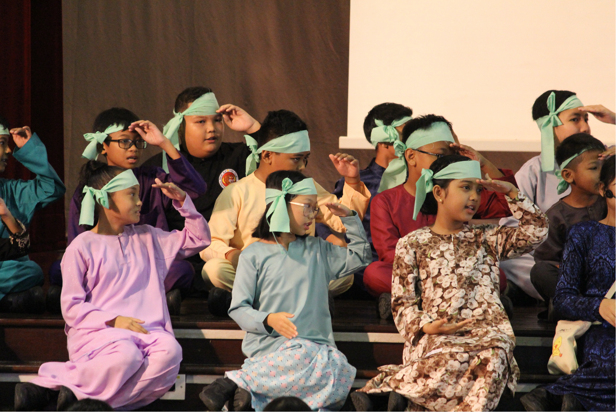 Children in colorful outfits wear green headbands and raise hands to brows, seated on stage steps.