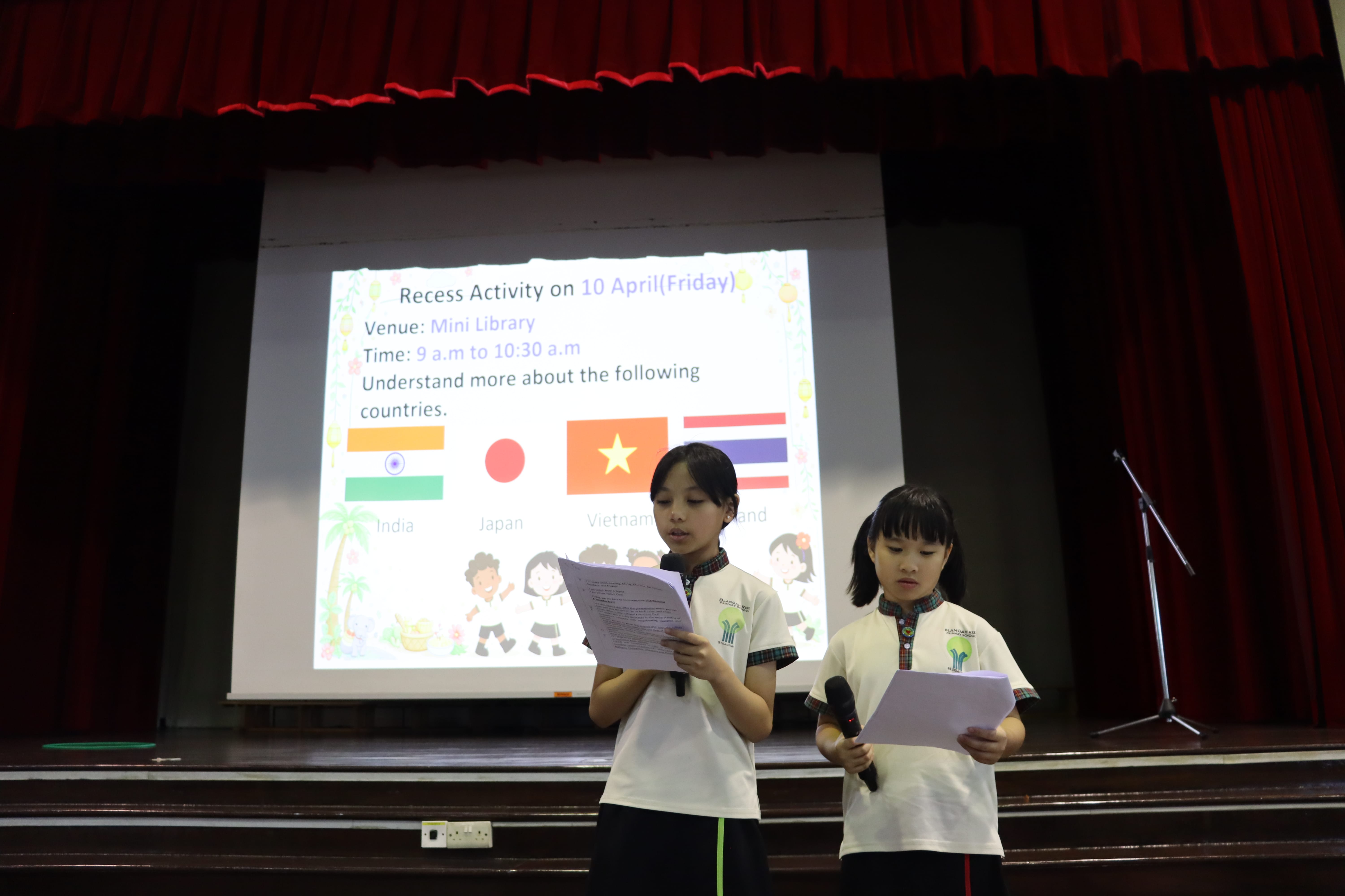 Two schoolgirls on stage read from papers; a projector behind shows flags and Recess Activity details.