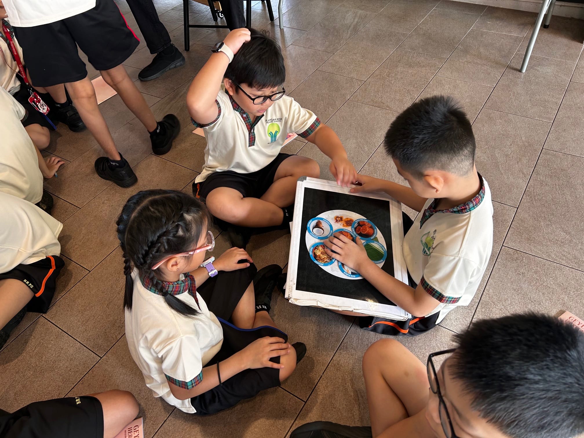 Three school children in uniforms sit on a tiled floor examining a board displaying a photo of a food plate.
