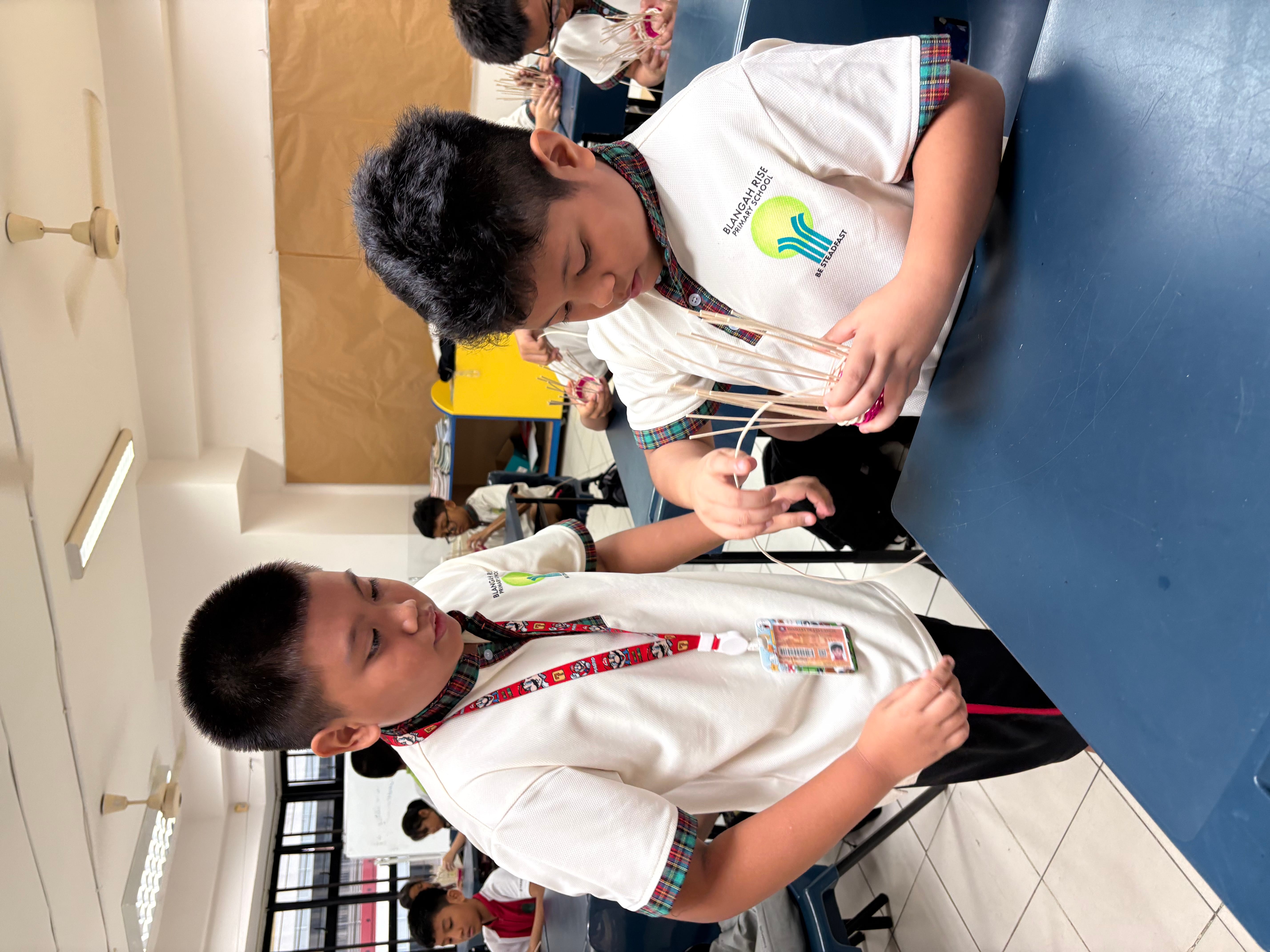 Two schoolboys in uniform build a spaghetti tower at a classroom table while others work in background.