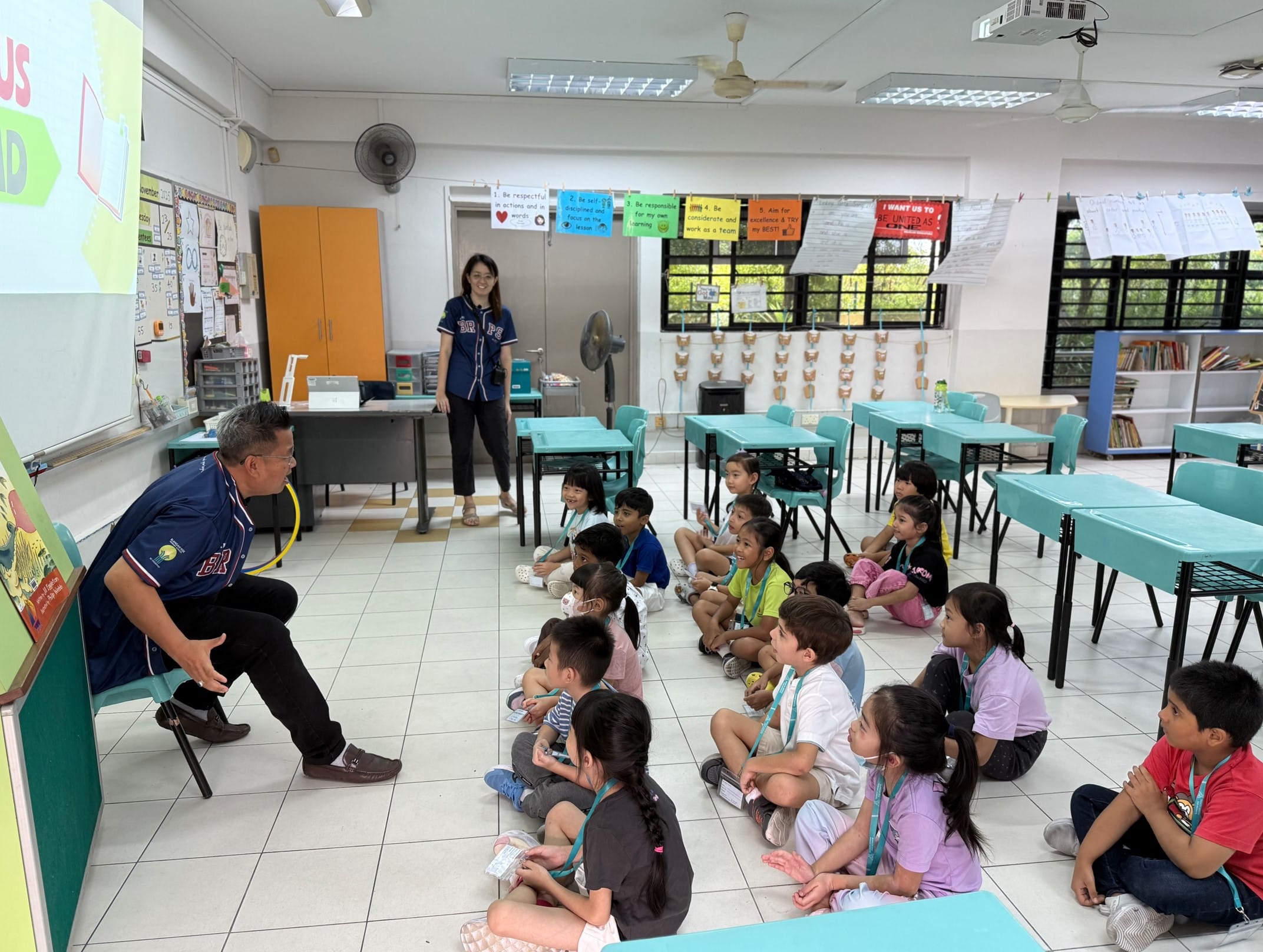 A man reads to young children sitting on a classroom floor while a woman stands nearby watching.