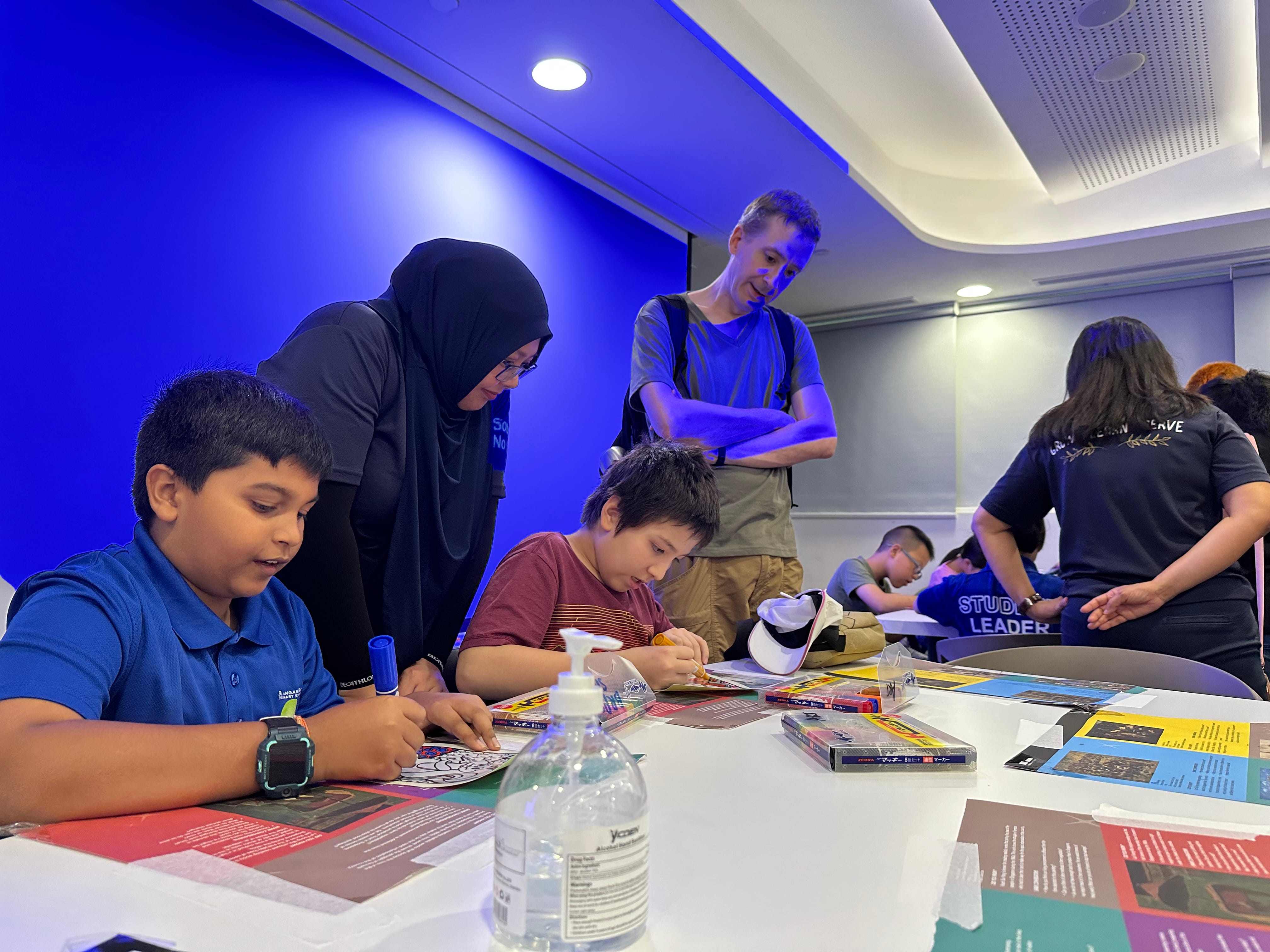 Children sit at a table doing a craft activity while adults supervise in a room with a bright blue wall.