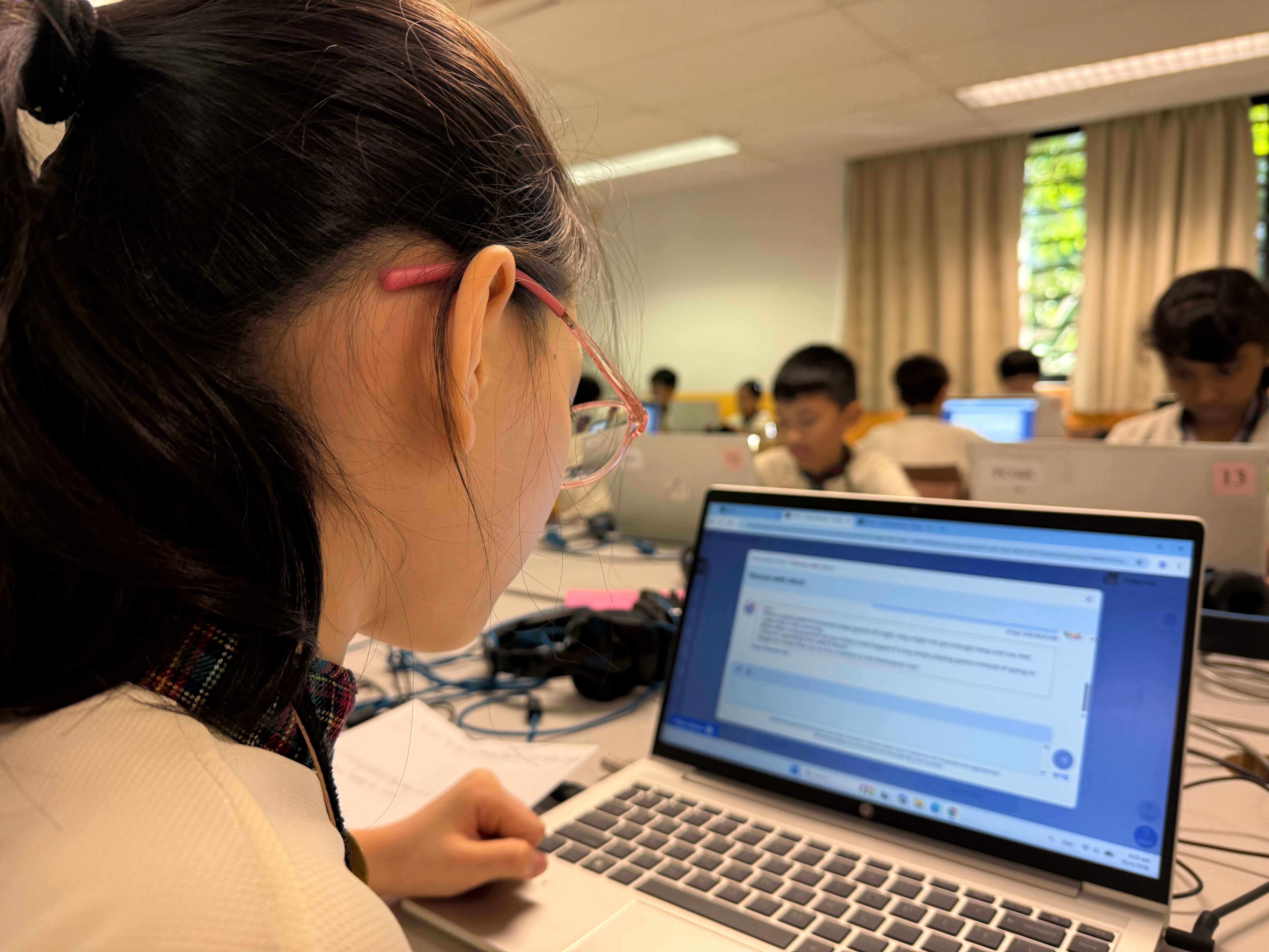 Student in glasses looks at a laptop in a classroom, with other students on computers in the background.