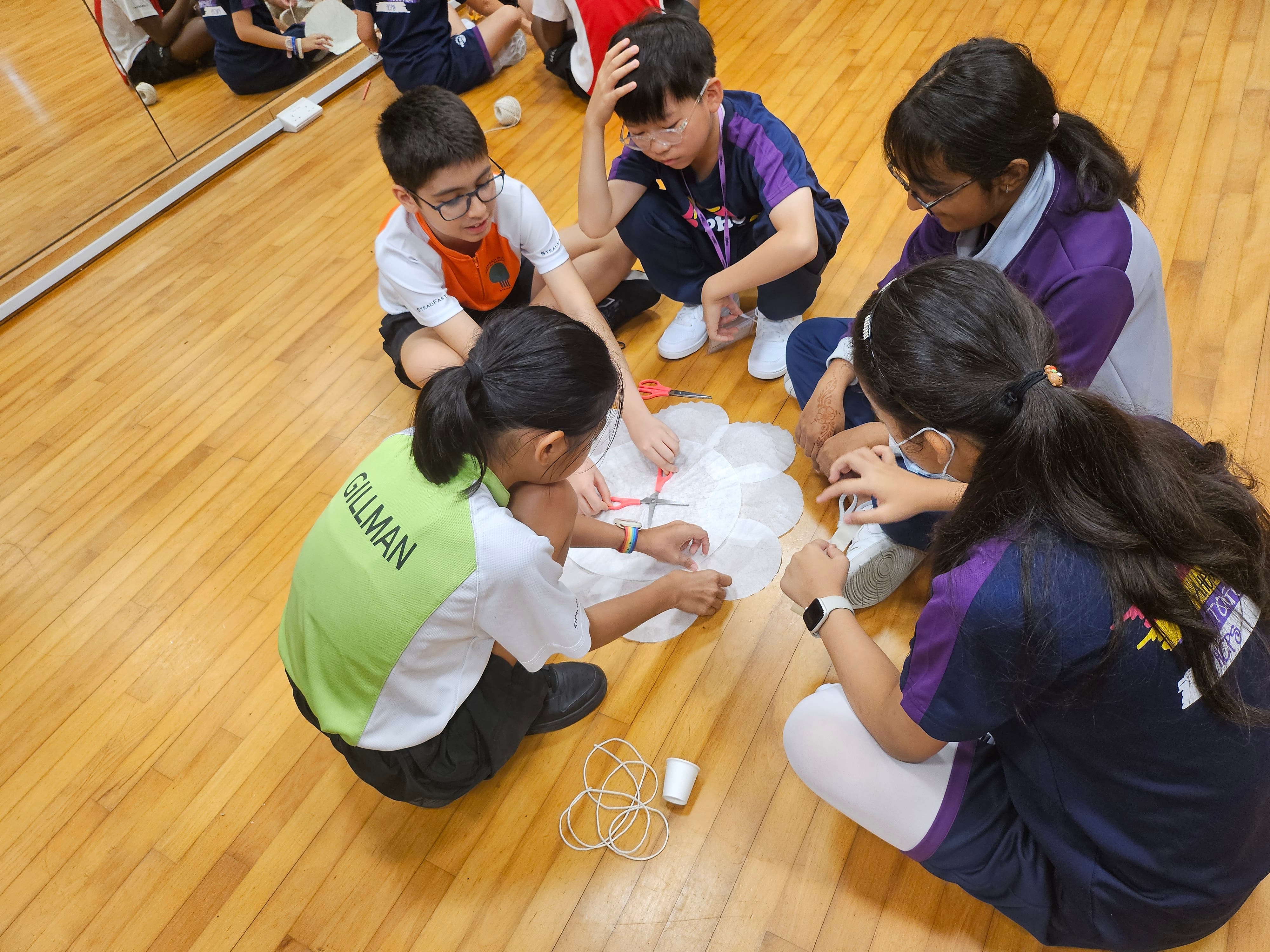 Five students sit on a gym floor, working together with paper circles, scissors, string, and a cup.