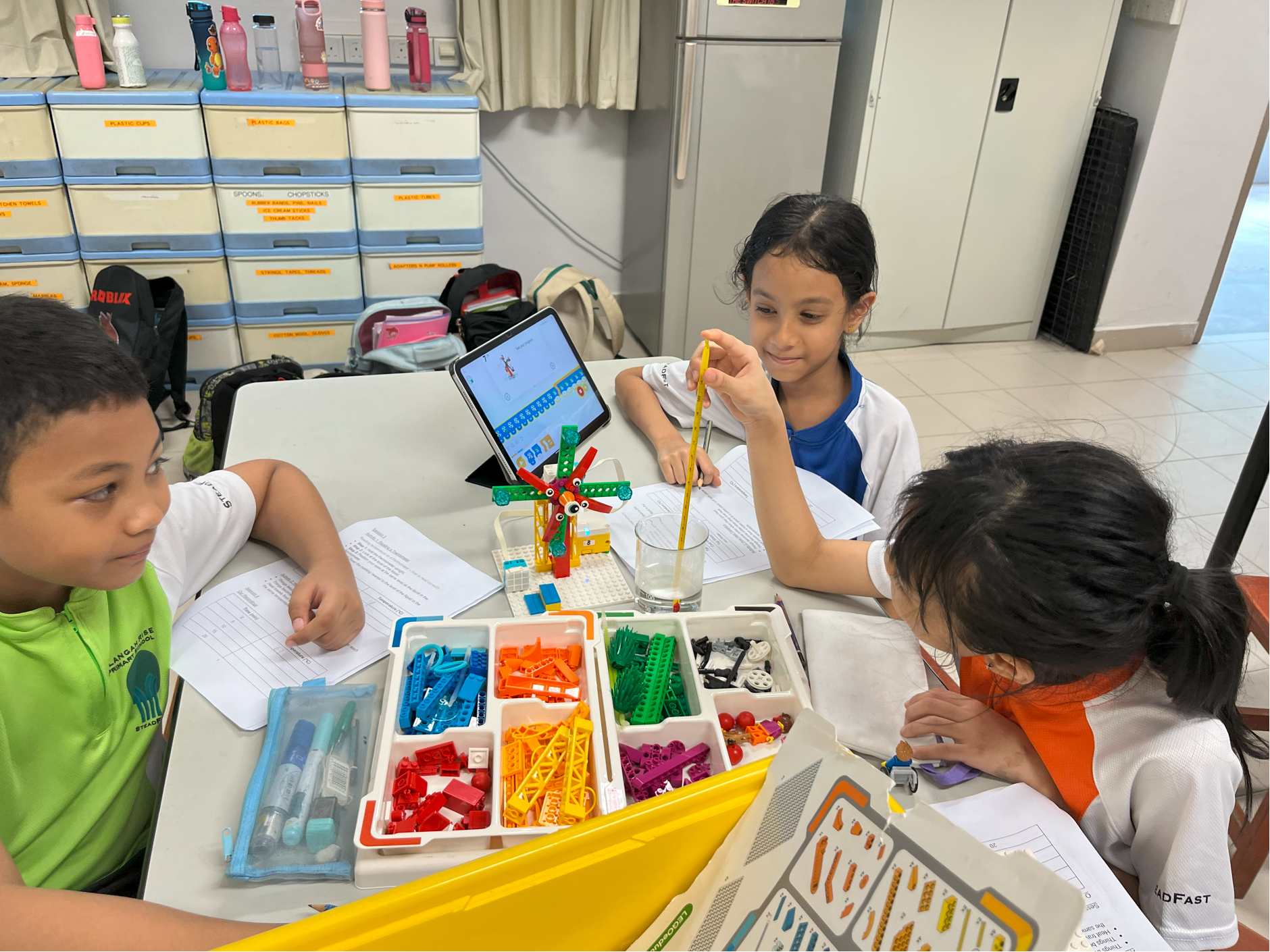 Three children at a table build a colorful LEGO model while one dips a straw in a glass; tablet on stand nearby.