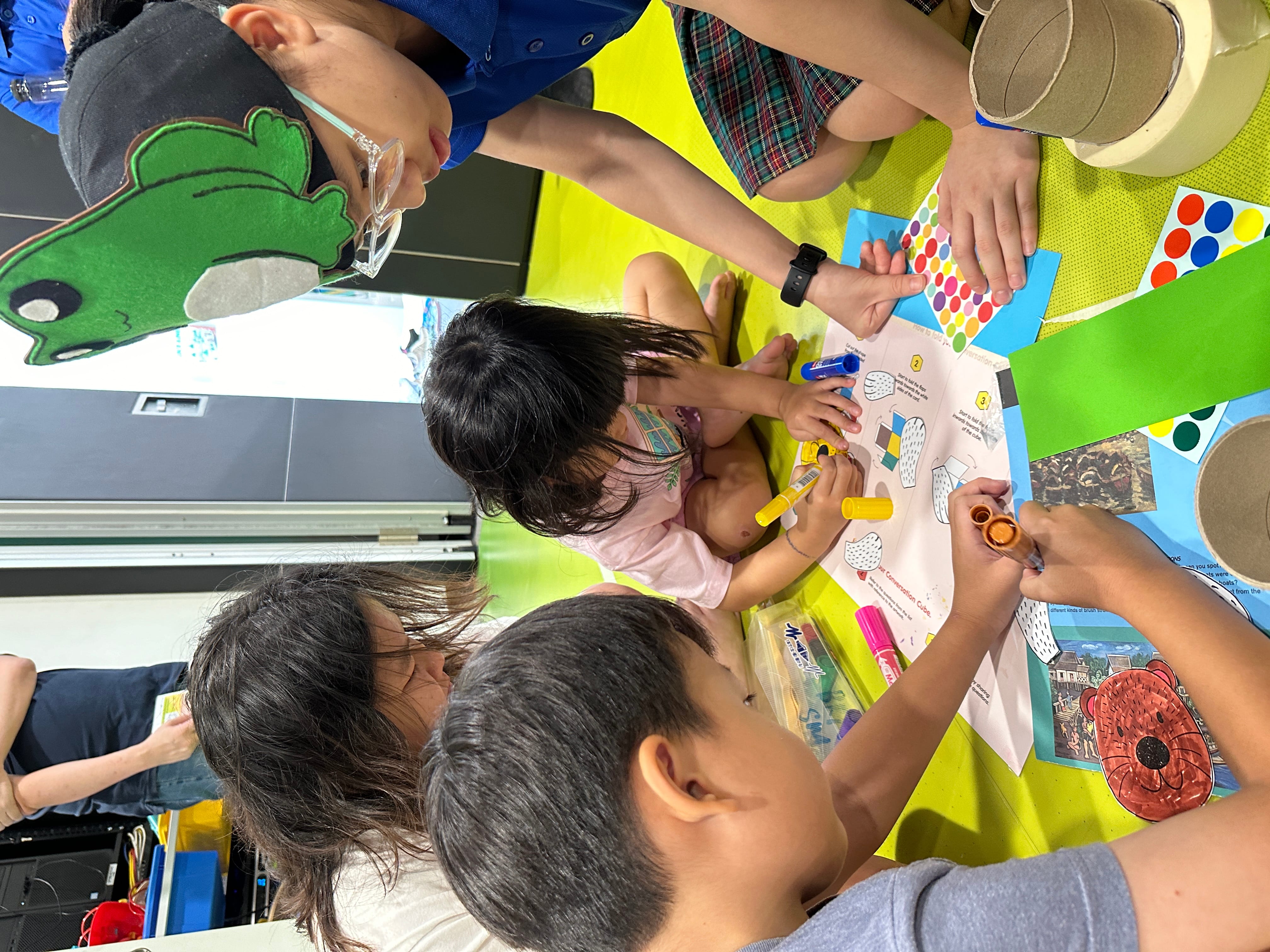 Four children sit at a table coloring a paper craft with markers, stickers, and cardboard tubes.
