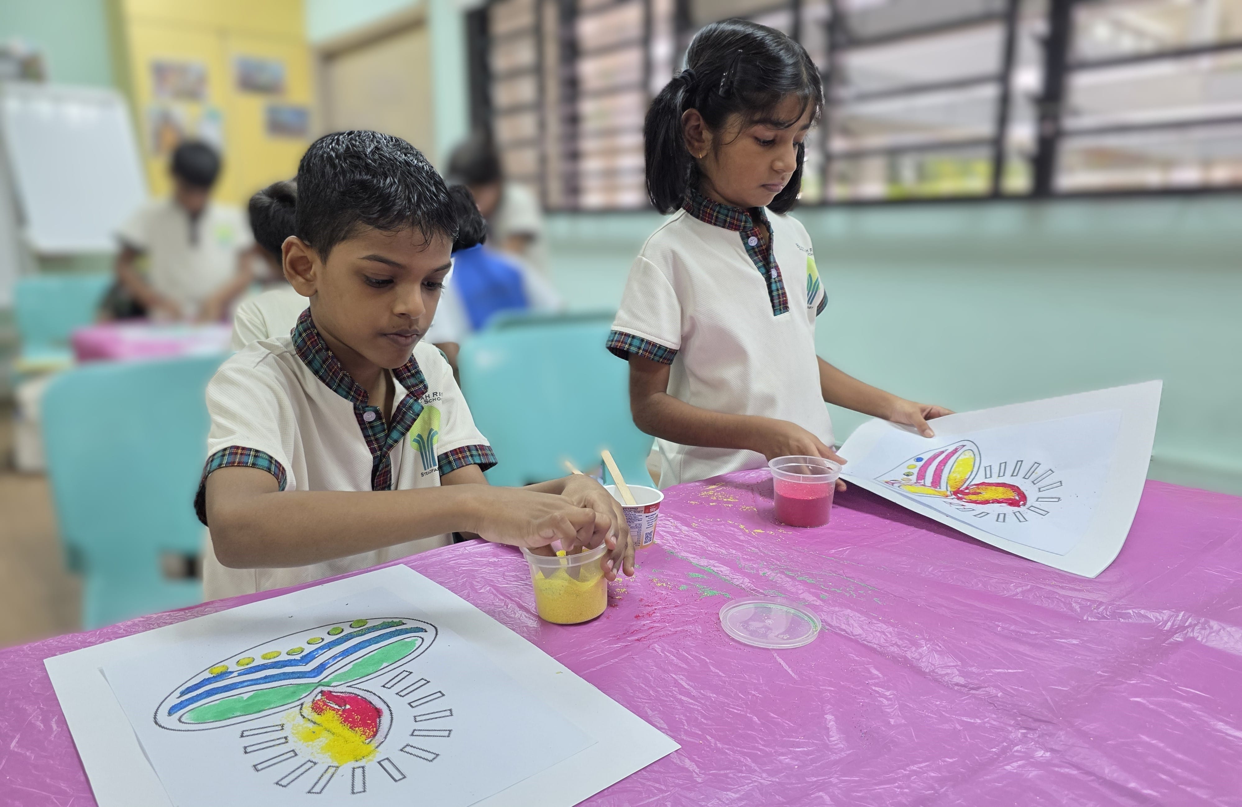 Two children in school uniforms color drawings at a table with cups of colored sand and a pink tablecloth.