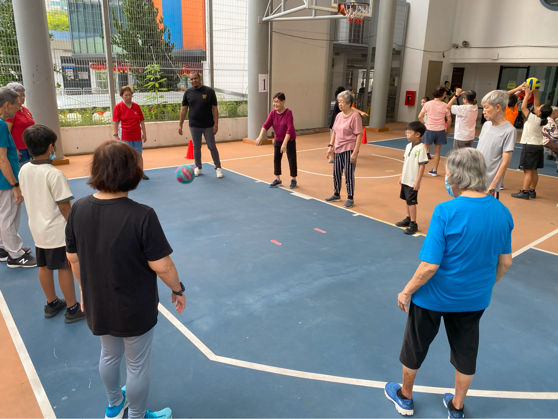 Group of seniors and kids stand in a circle on an indoor court, watching a colorful ball roll.