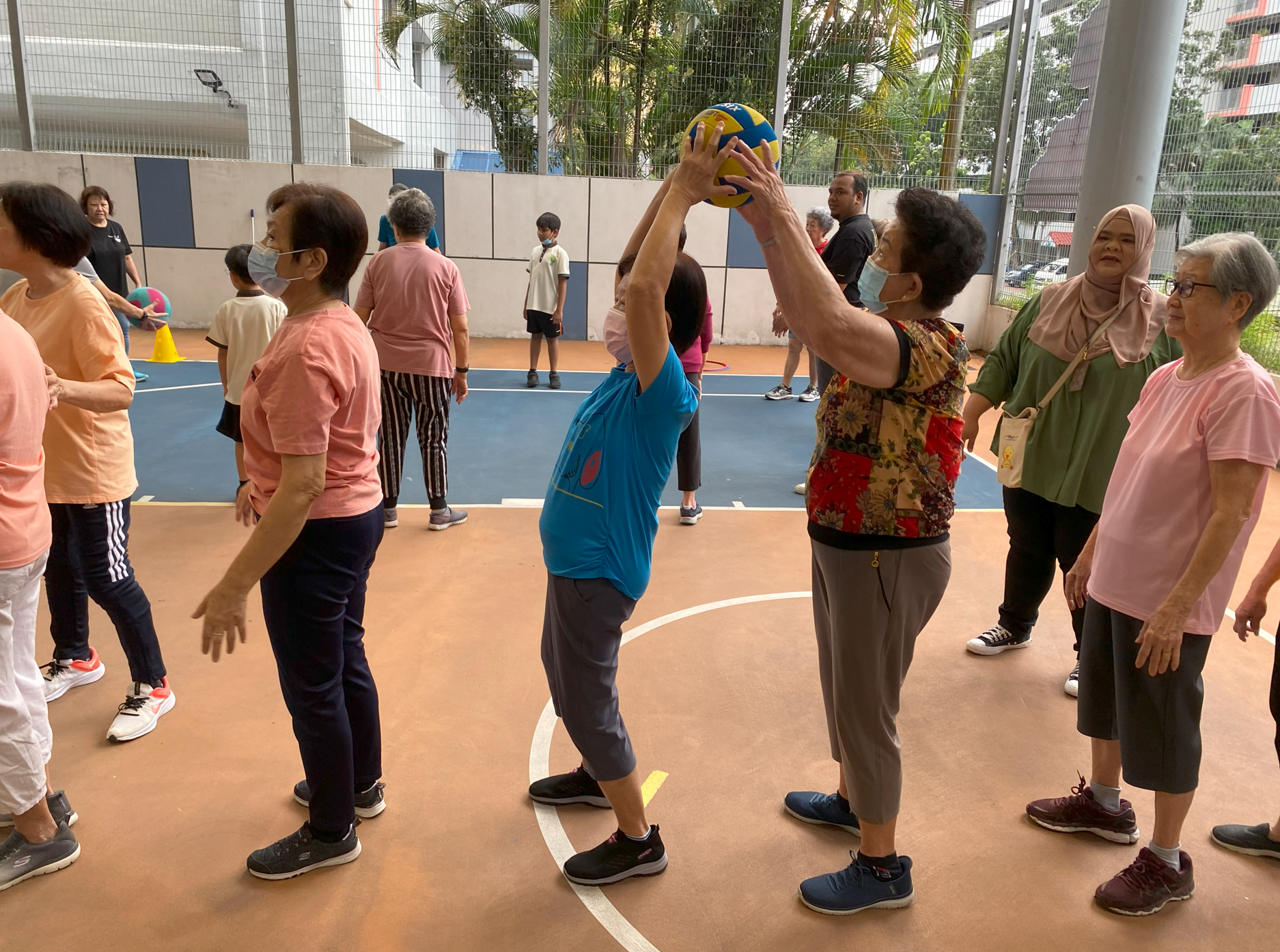 Group of older adults on a court; one in blue raises a volleyball overhead while others watch nearby.
