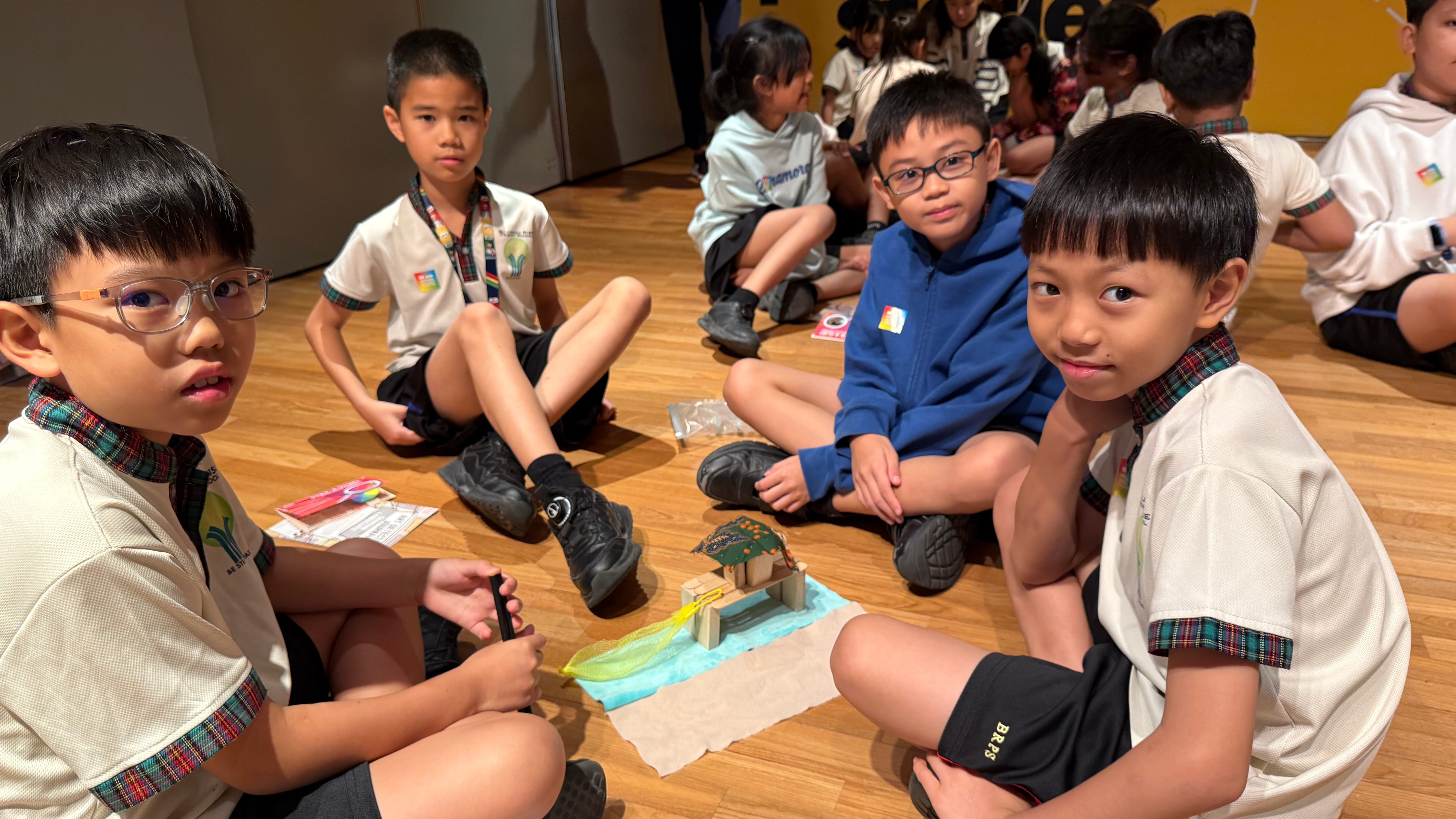 Four schoolboys sit on a wooden floor around a small craft model; more children sit in the background.