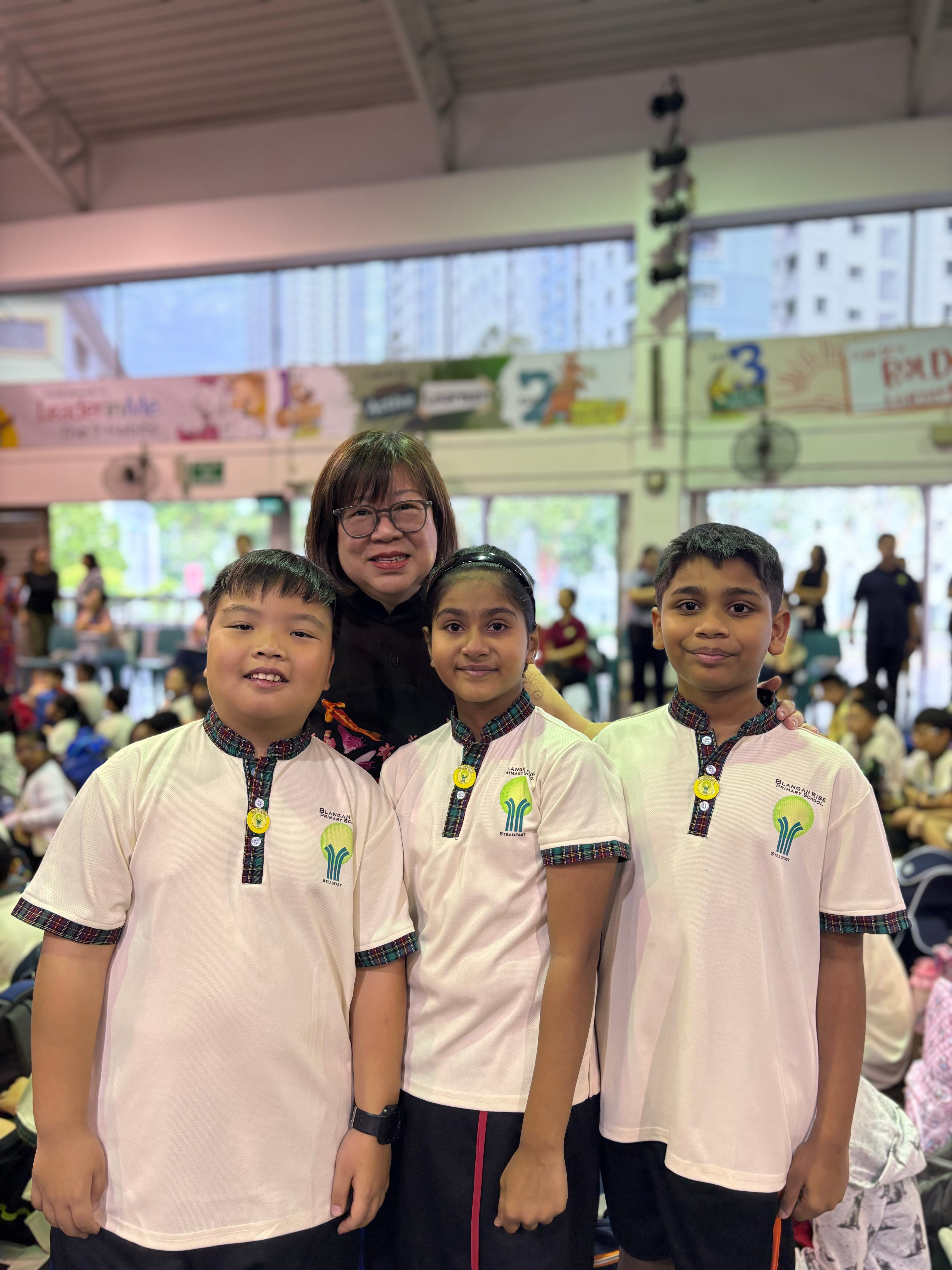 An adult woman with glasses poses with three children in matching white school uniforms inside a busy hall.