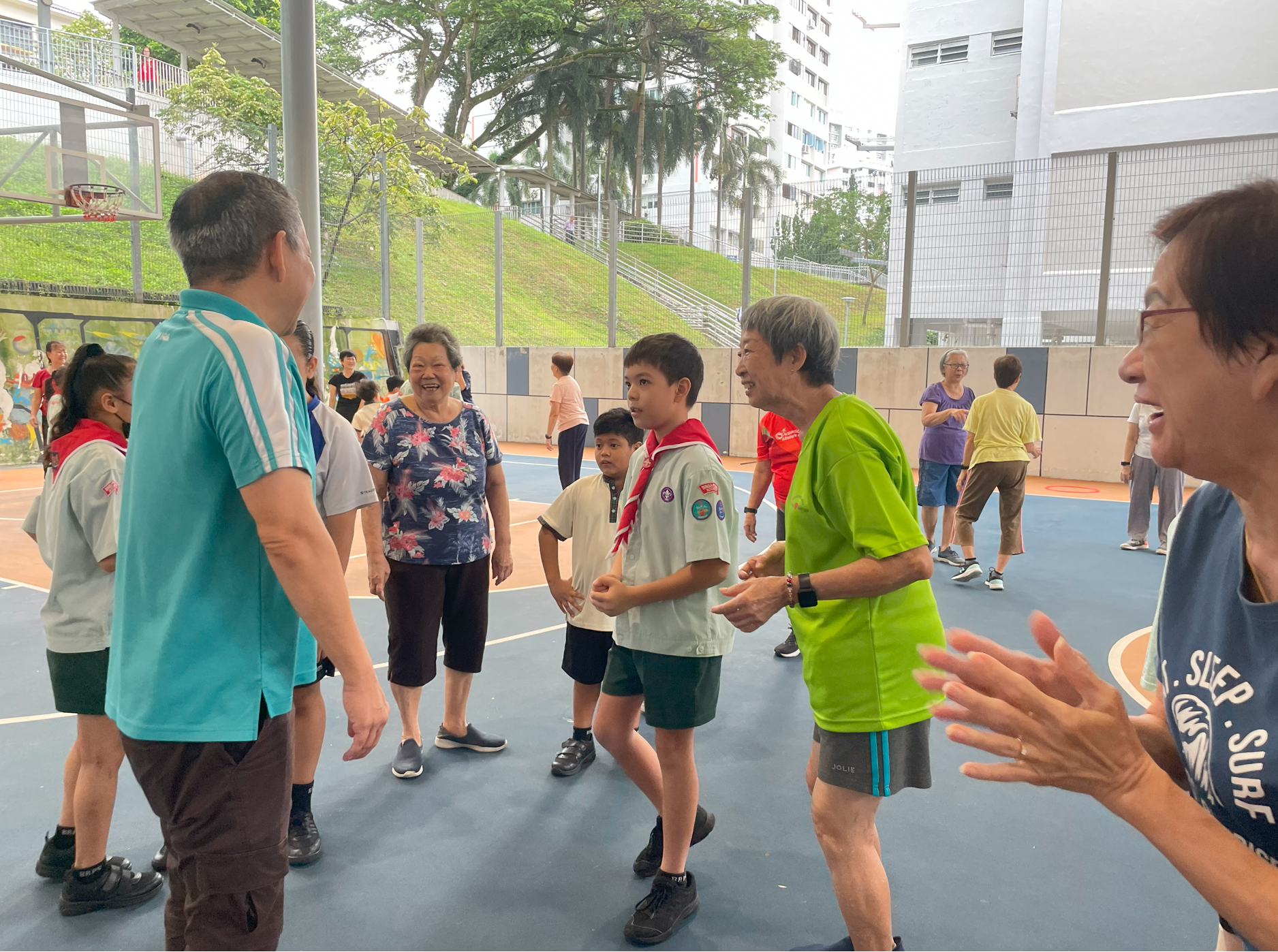 Group of adults and children chatting and laughing on an outdoor court, with buildings and trees behind.