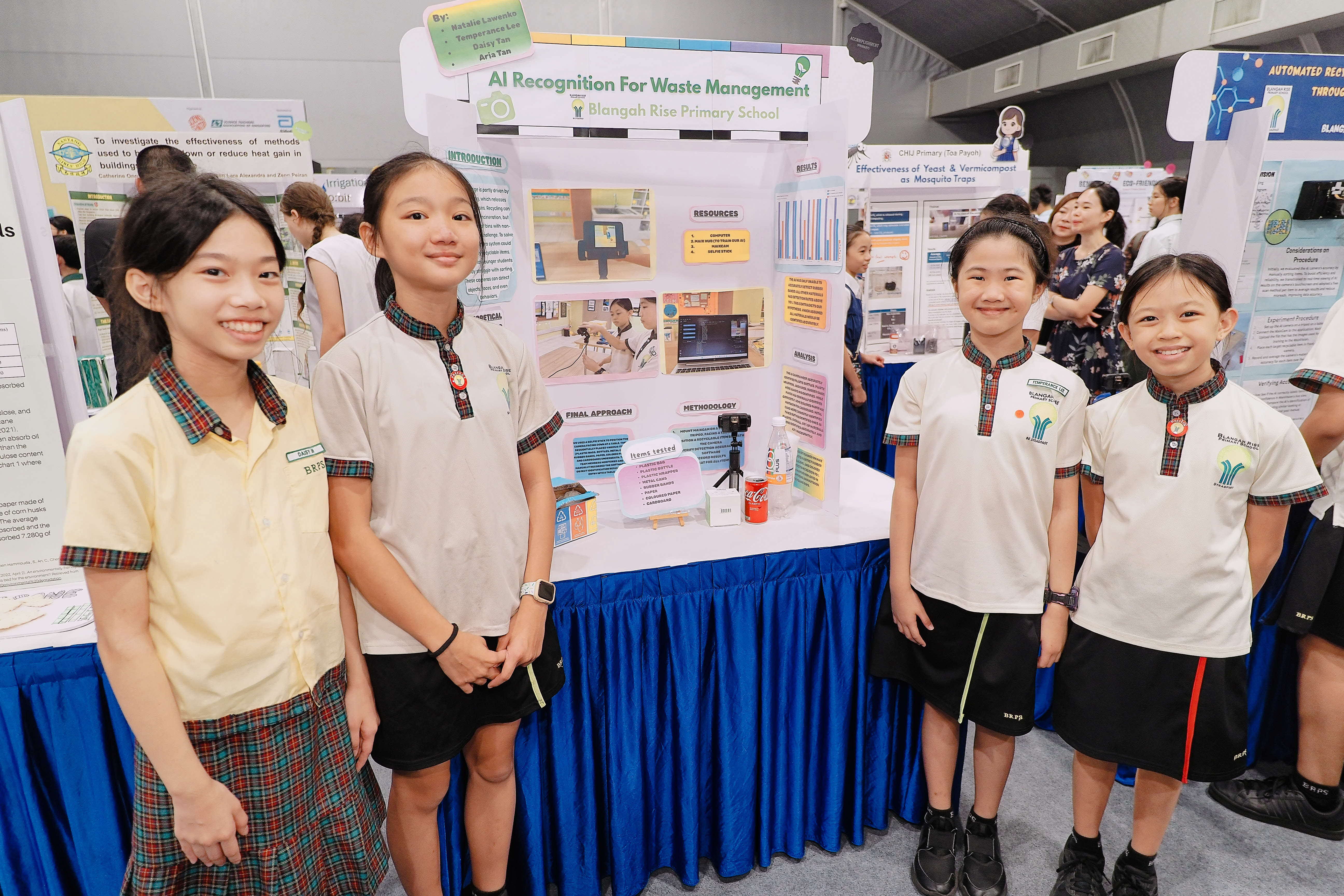 Four primary school students in uniform stand beside a science fair poster titled AI Recognition For Waste Management.