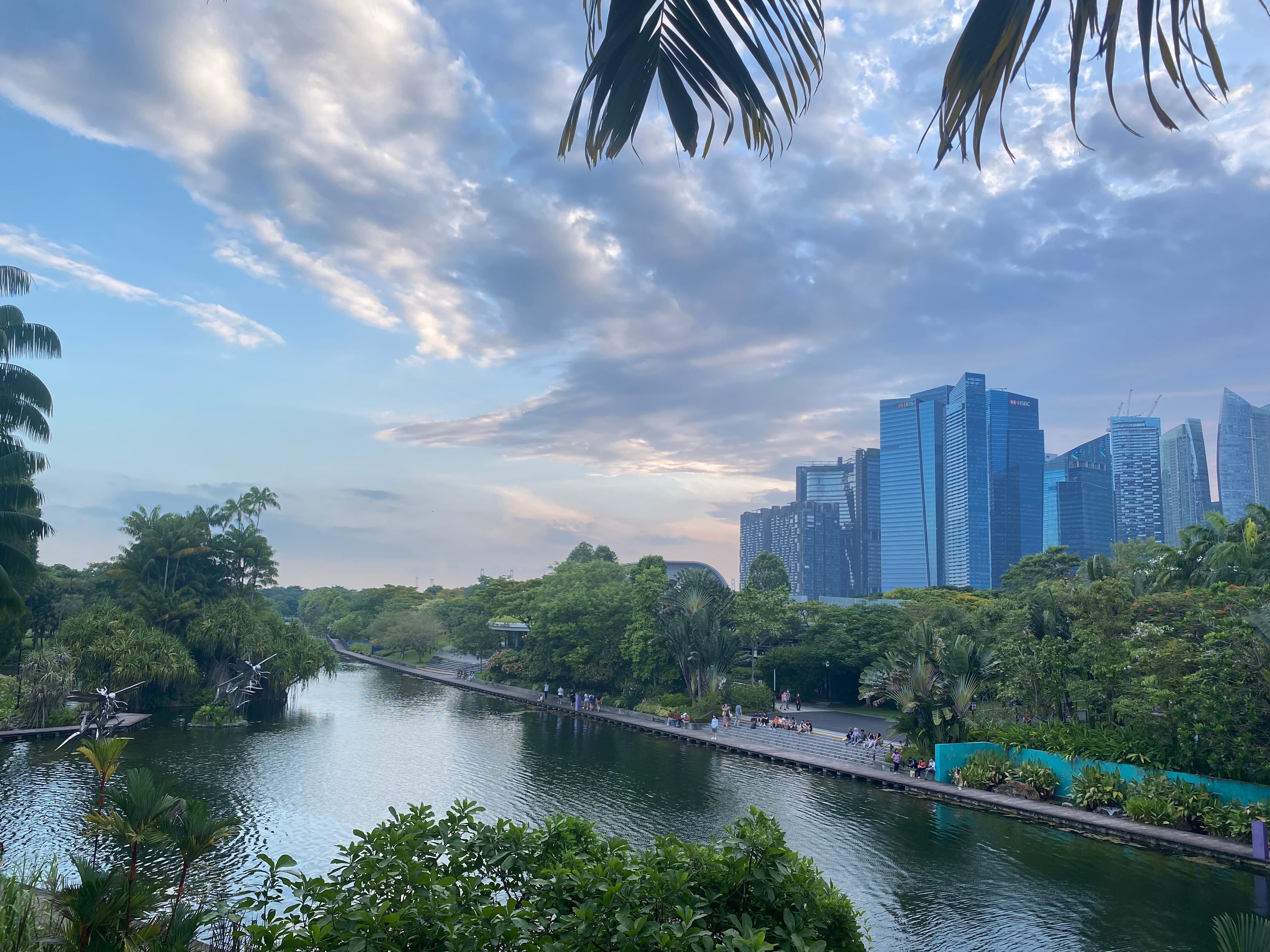 A calm urban waterway lined with lush greenery and a walkway, with tall glass skyscrapers under a cloudy sky.