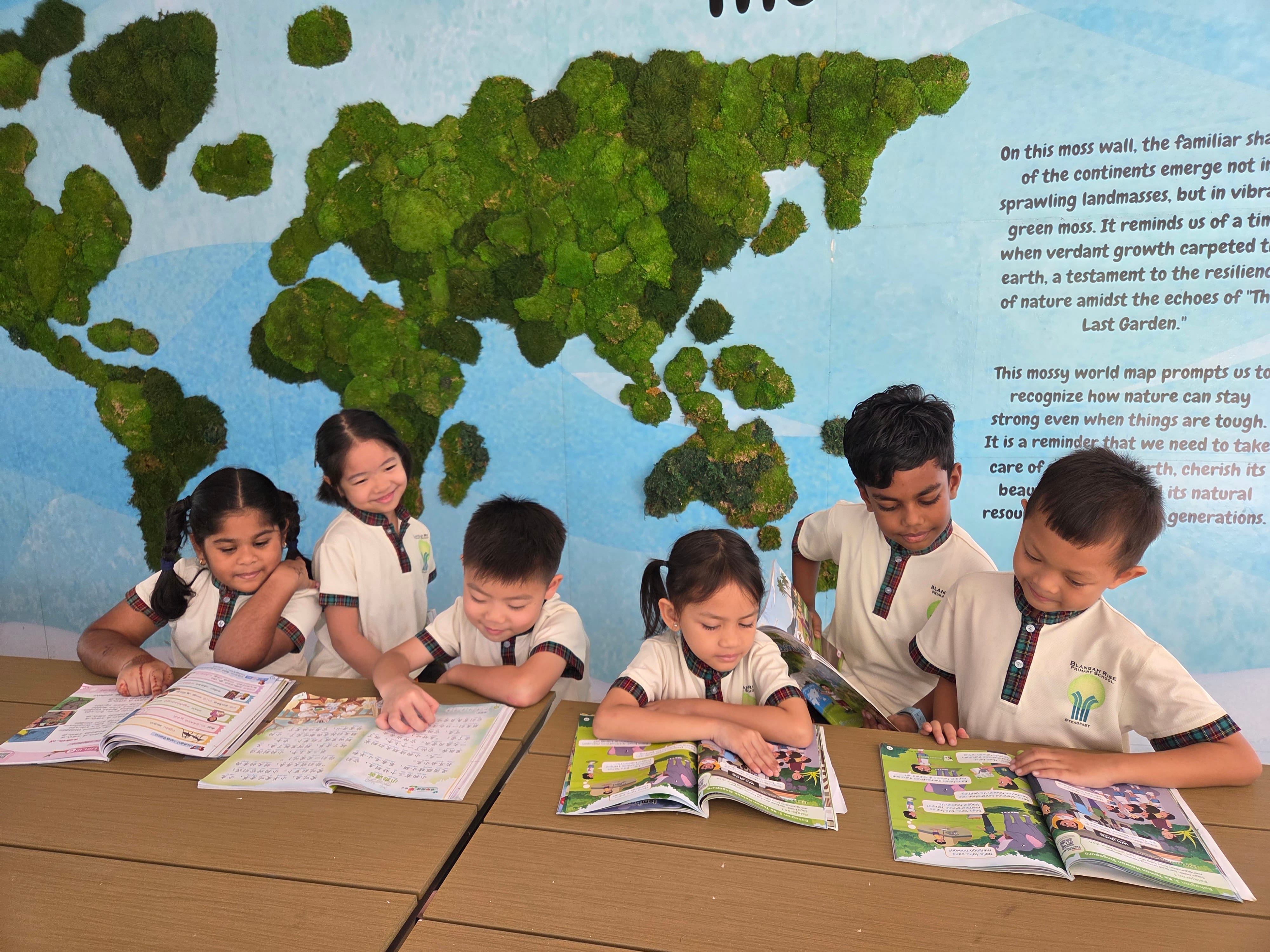 Six young students in white uniforms read open books at a table in front of a green moss world map wall.