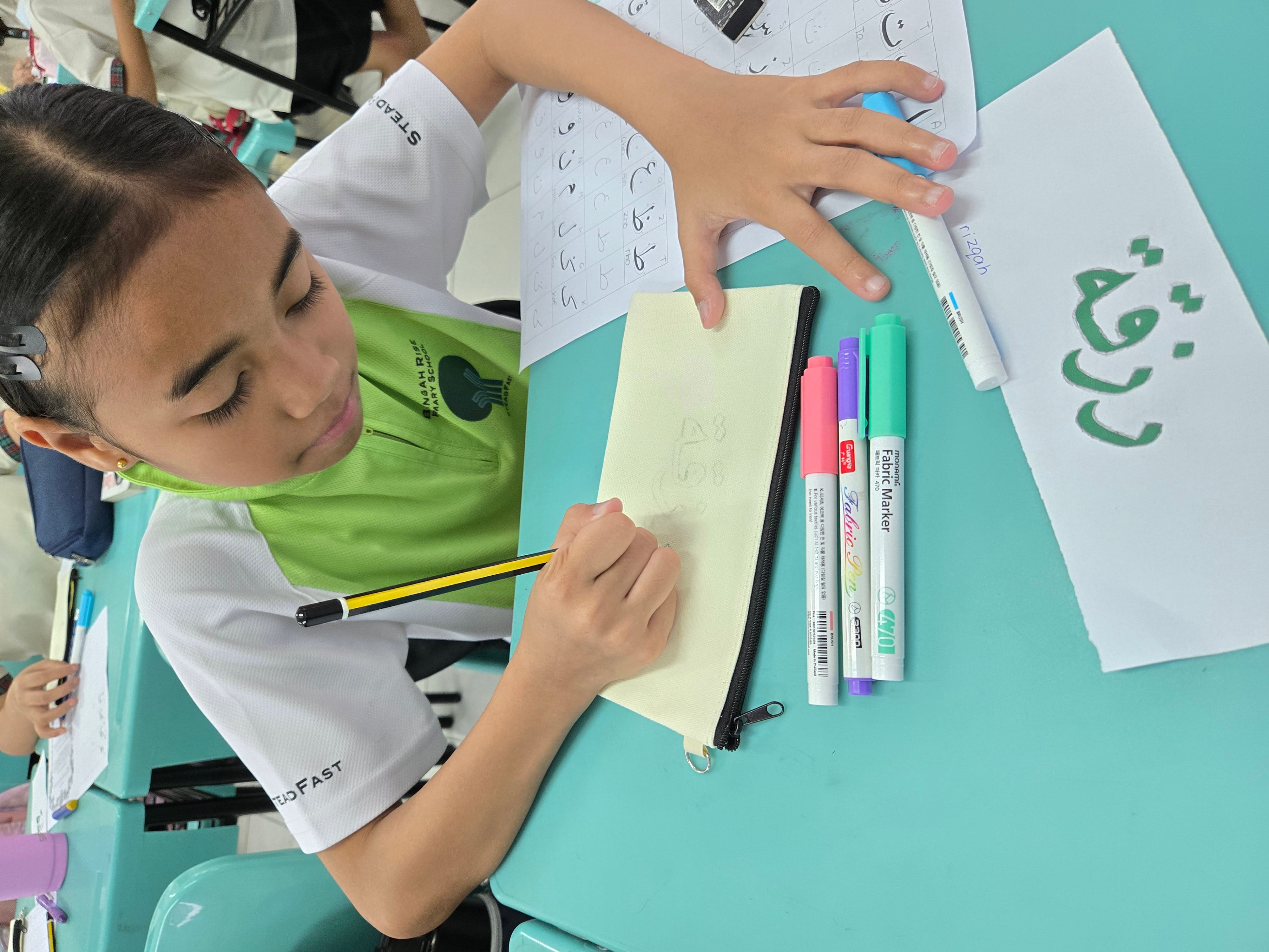 A young student in a school uniform writes on a pencil case at a teal desk with fabric markers nearby.