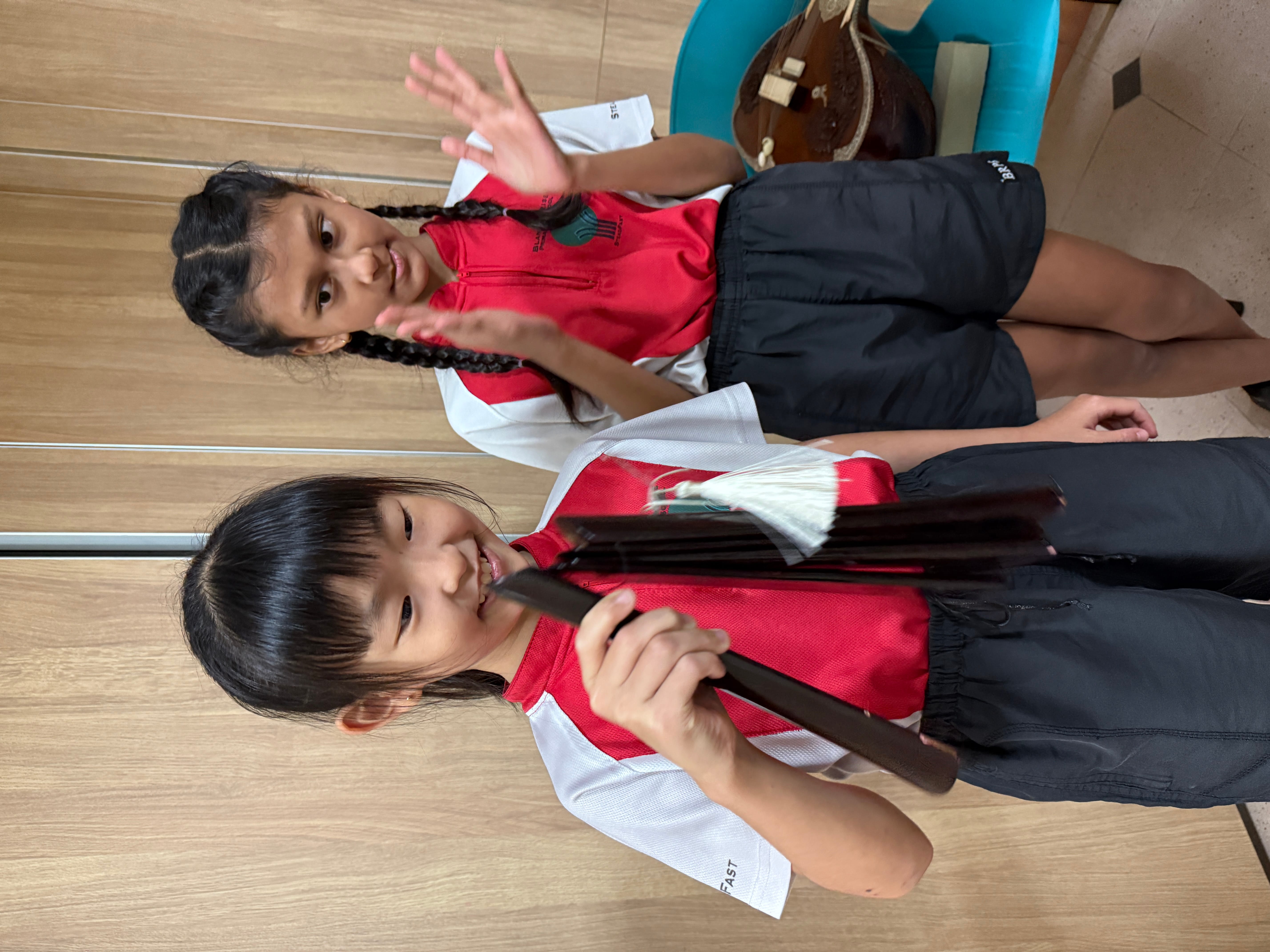 Two children in red-and-white shirts, one holding chimes, the other clapping, indoors by wooden wall.